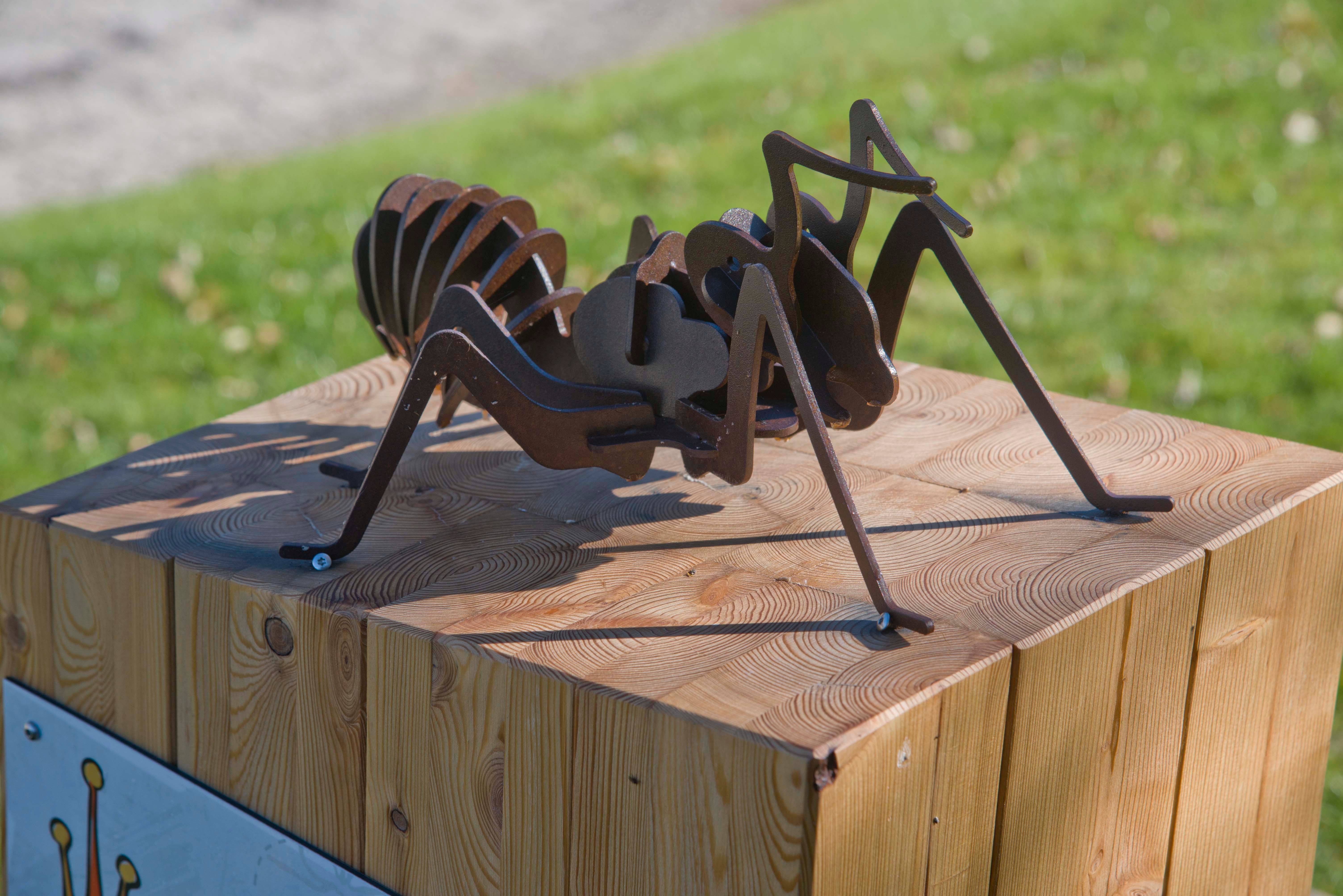 Metal sculpture of an ant on a wooden block outdoors.