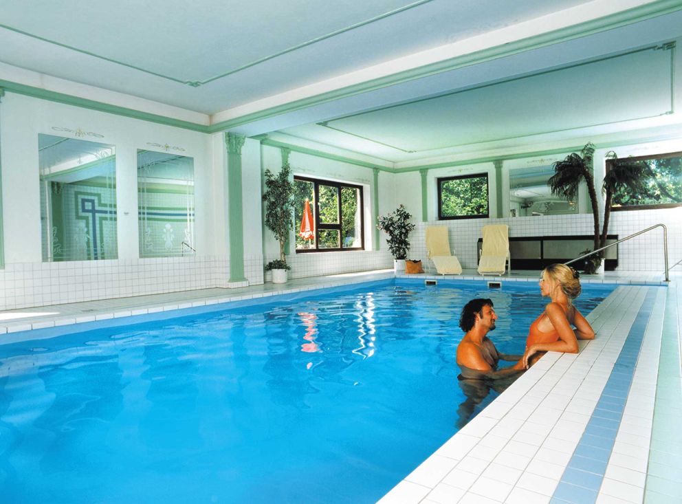 A couple in the pool of an elegant indoor area with plants and sun loungers.