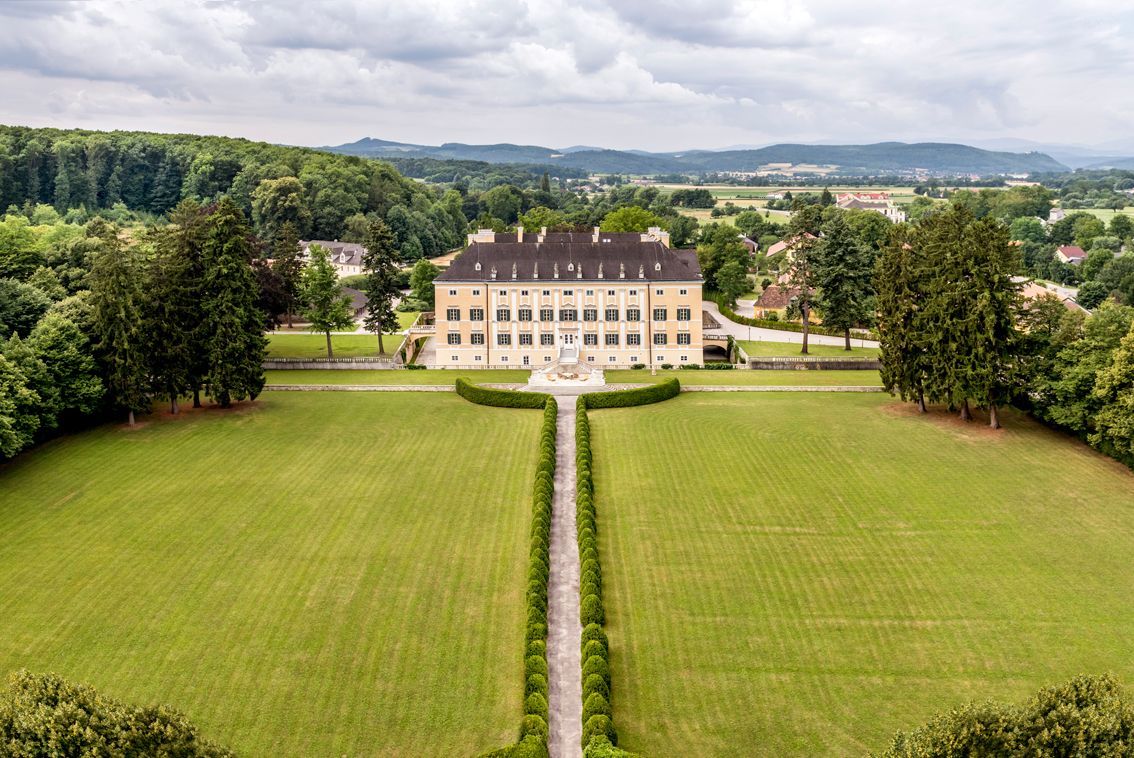 Aerial view of Frohsdorf Castle, a large, symmetrical castle with a yellow façade, manicured lawn and trees in the background.