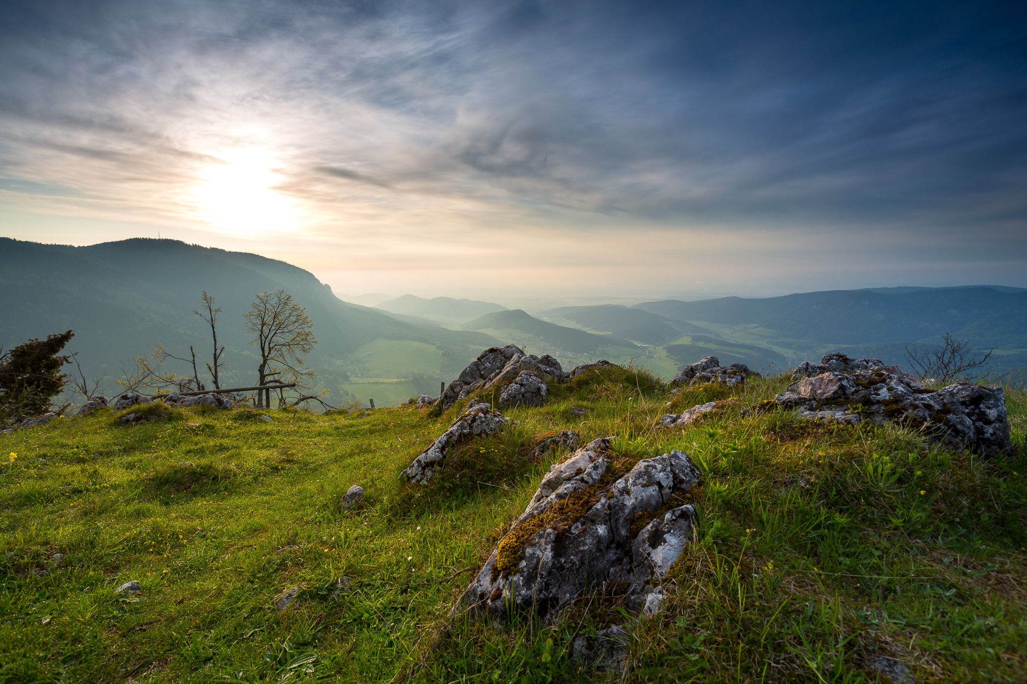 Panoramic view from a hill with rocks and grass in the foreground, mountains and a cloudy sky in the background.