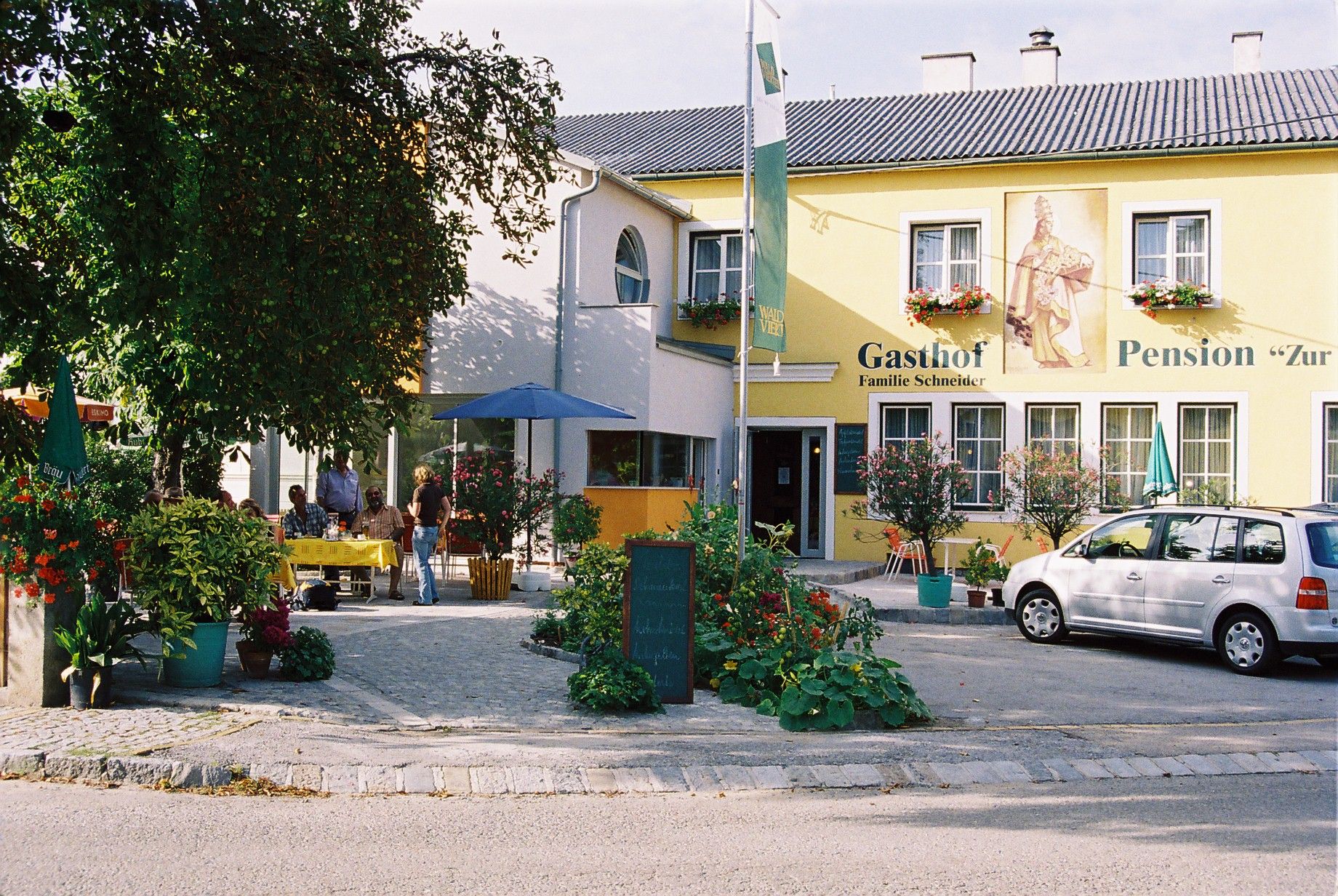 A yellow building with the inscription 'Gasthof-Pension Zur Hammerschmiede'. There are tables, chairs and a car in front of the building.