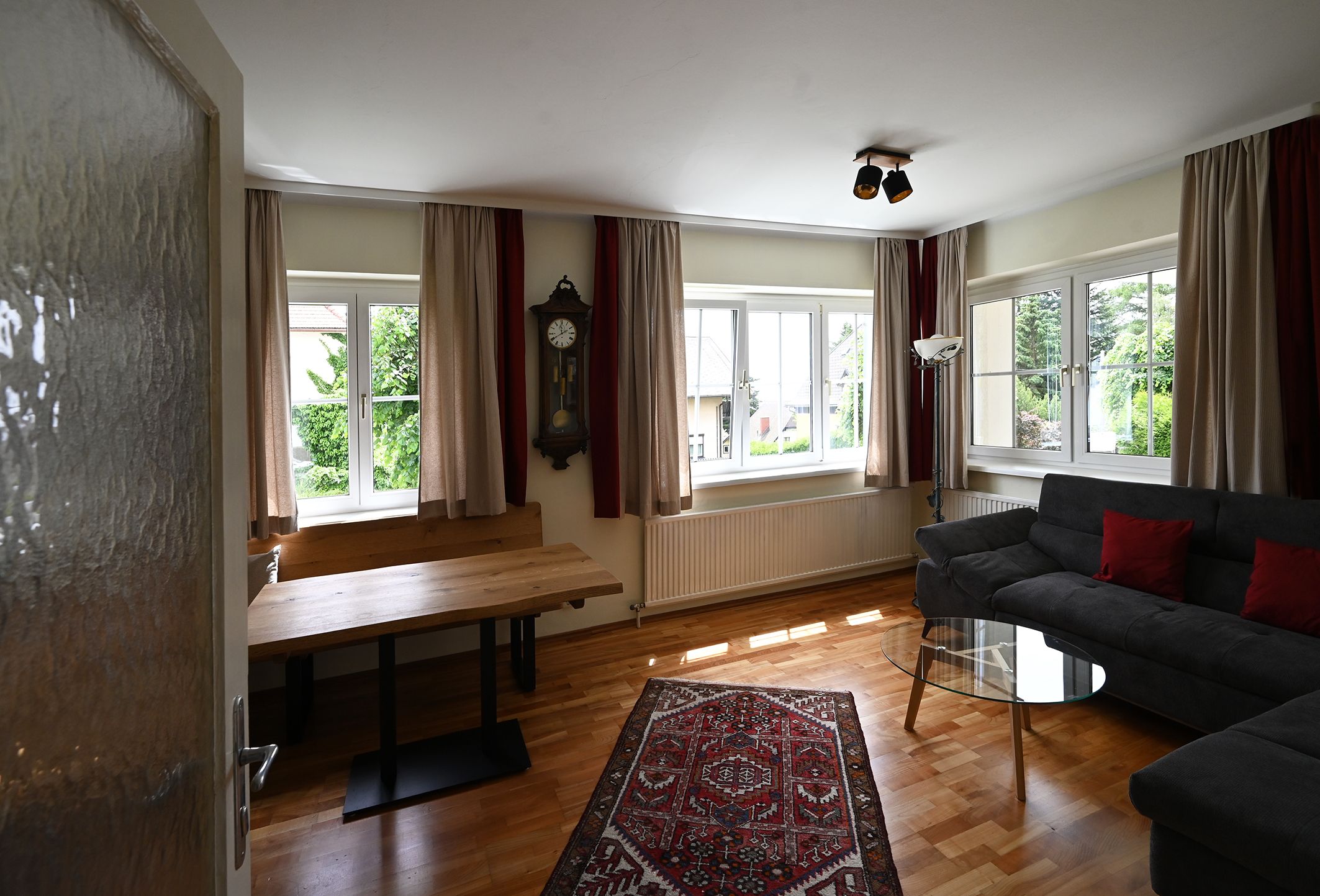 A living room with wooden floor, gray sofa, glass table, wall clock and carpet. Large windows with curtains let in plenty of light.