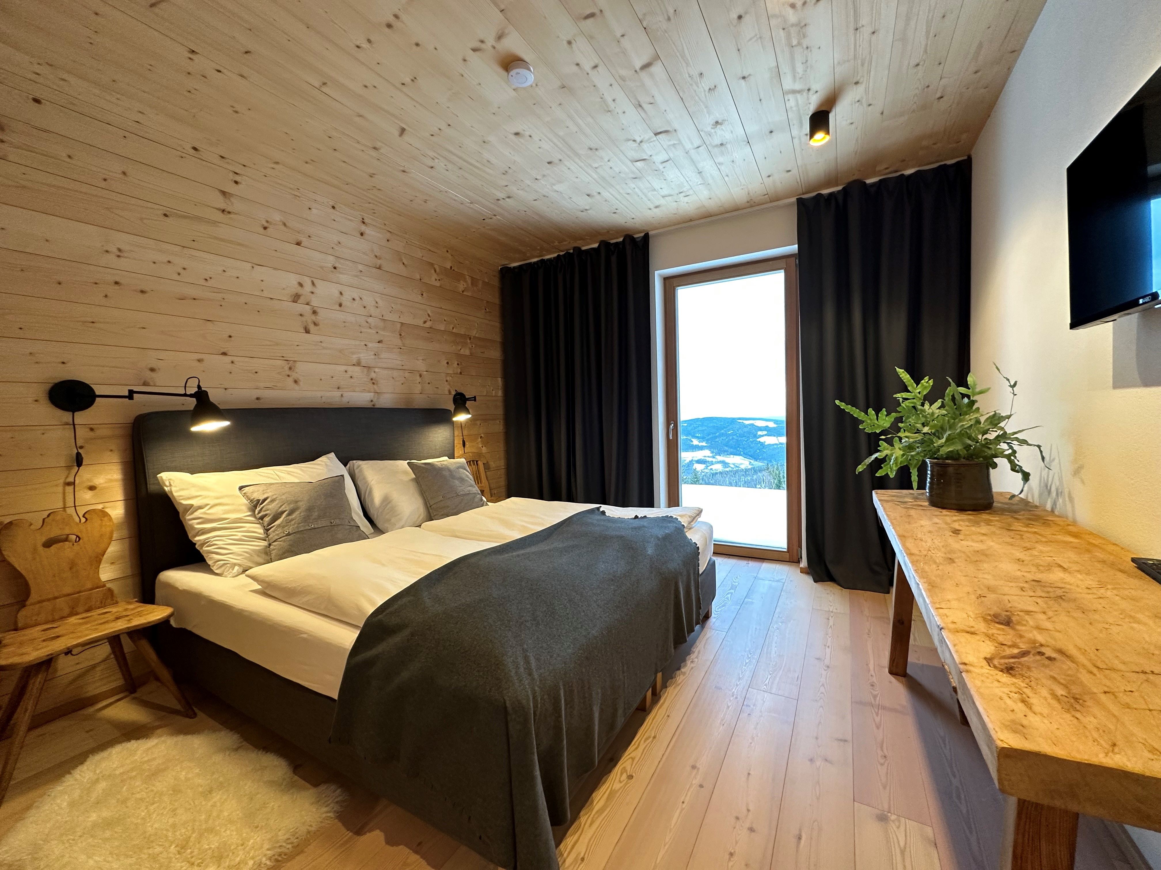 Cozy bedroom with wooden walls, double bed, wooden chair, table with plant and large window with curtains.