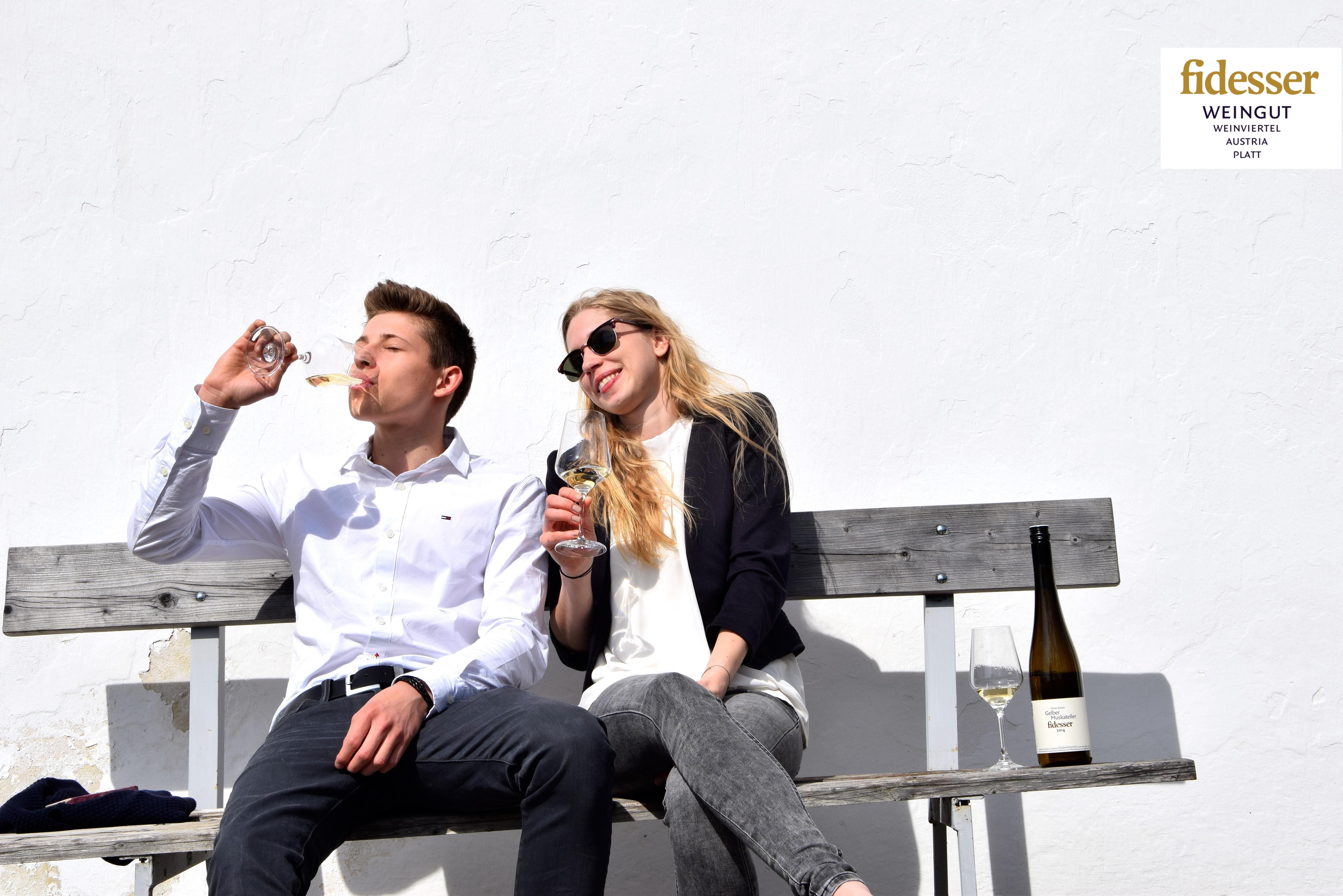 Two people are sitting on a bench drinking wine in front of a white wall.