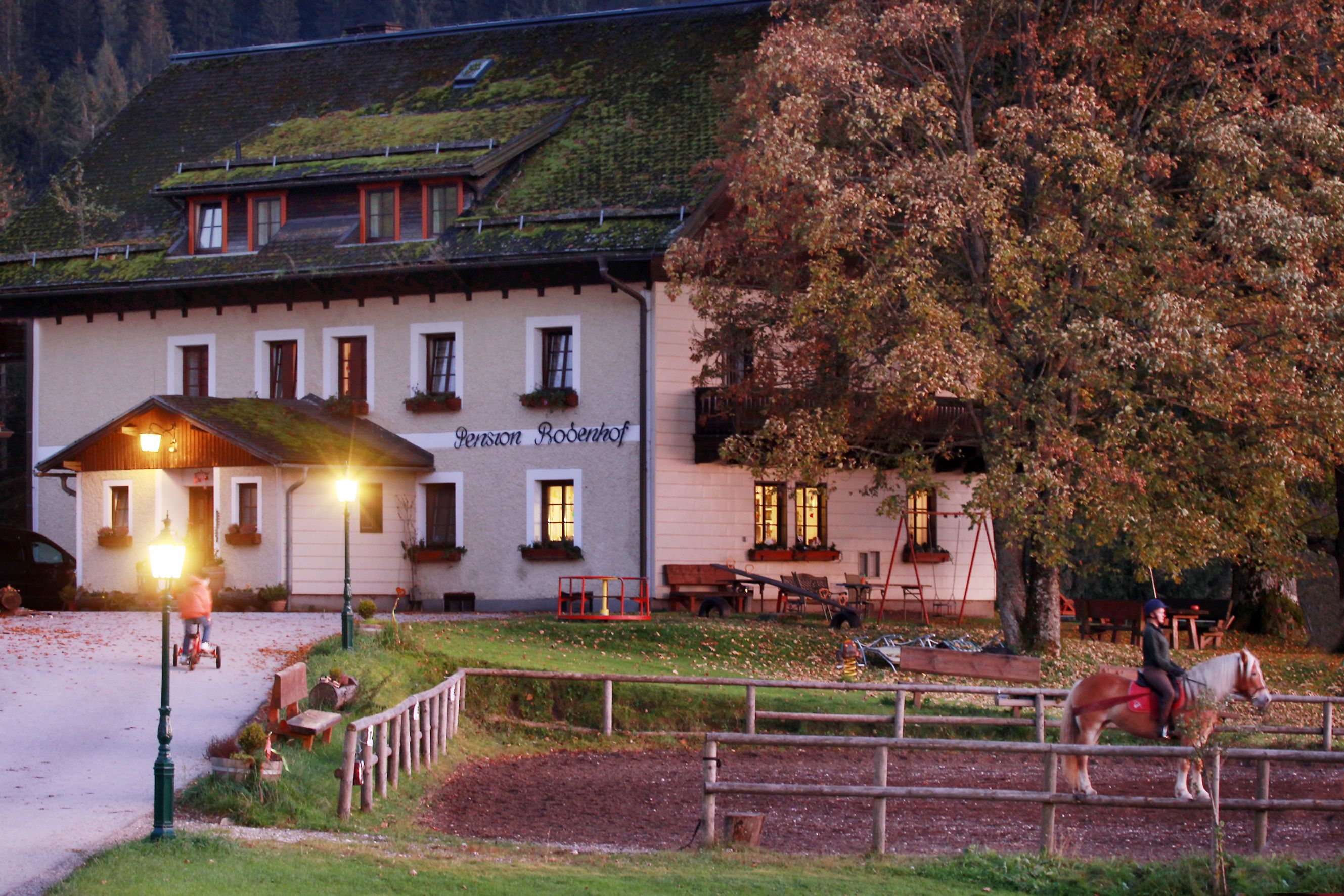 A farm with illuminated windows, a rider on a horse and a child on a tricycle in the foreground.