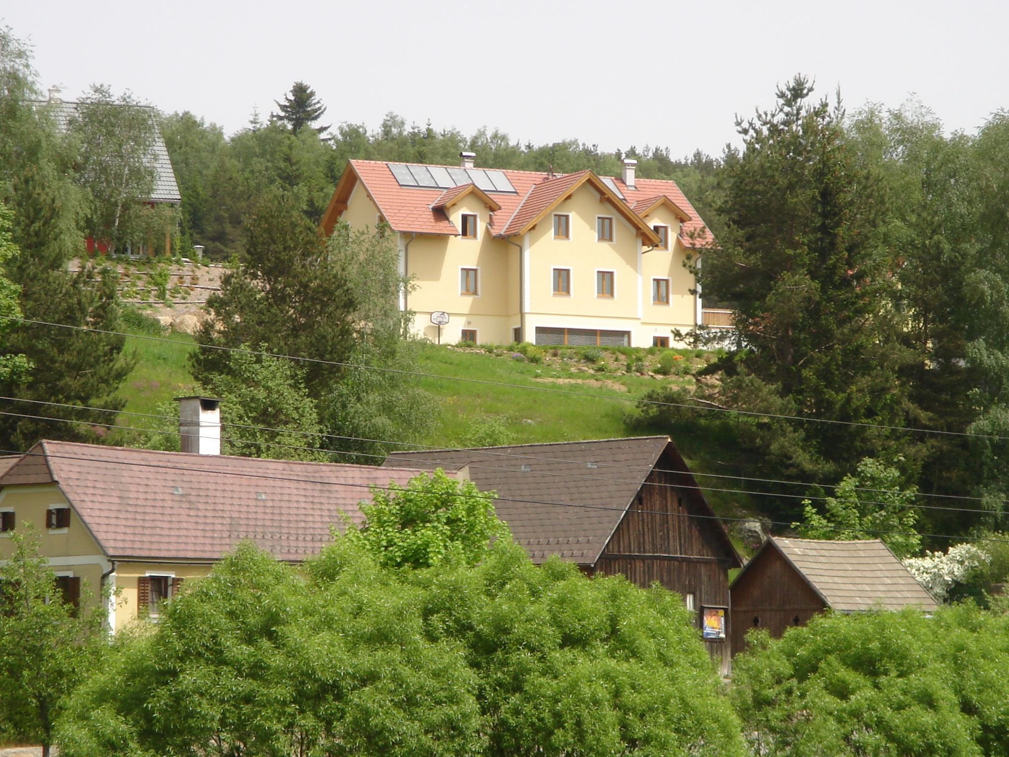 A large yellow house with solar panels on the roof stands on a hill, surrounded by trees and smaller buildings in the foreground.