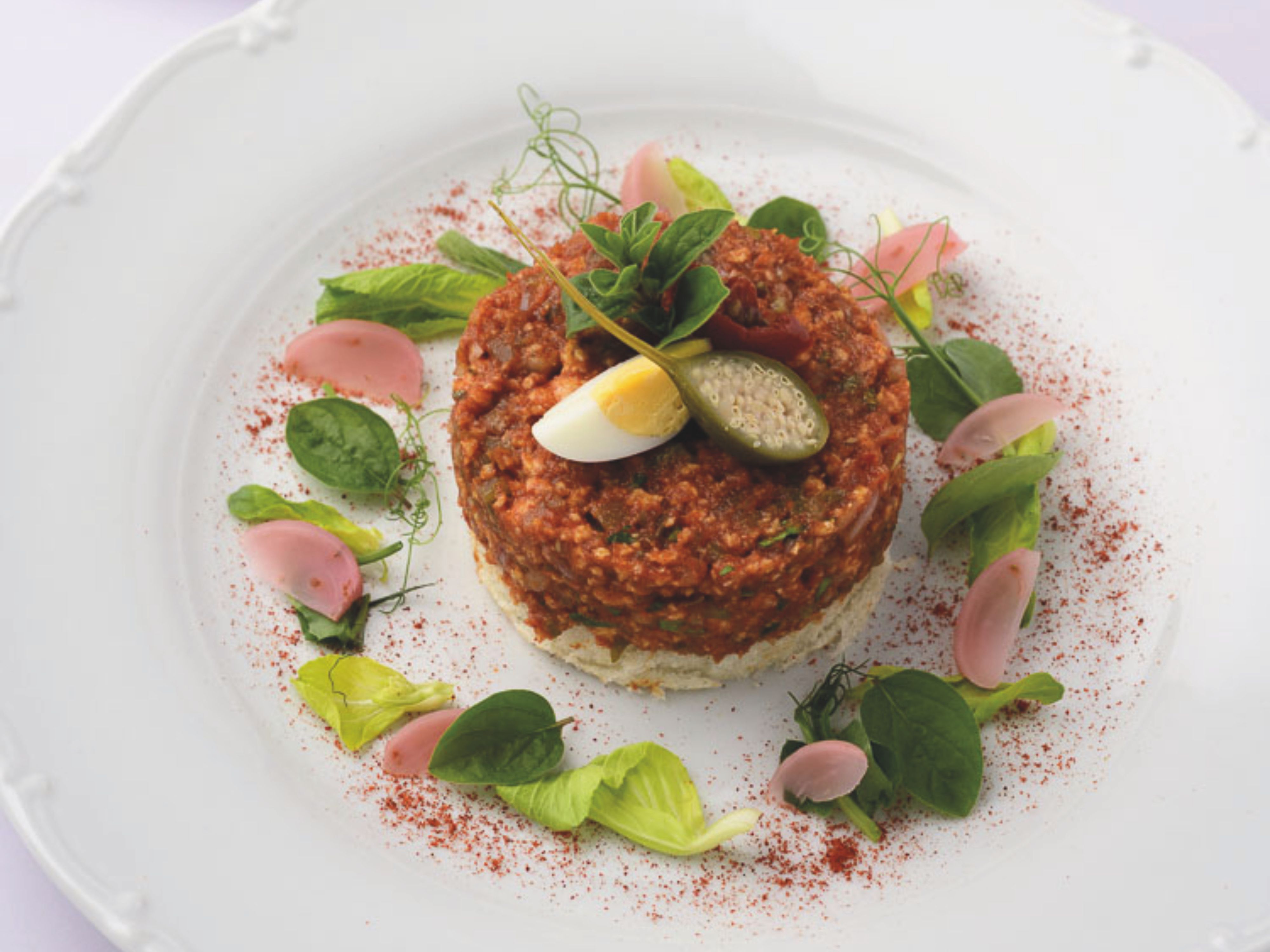 An artfully plated beef tartare on a white plate, consisting of a round portion of tartare garnished with capers, egg slices, and fresh herbs.