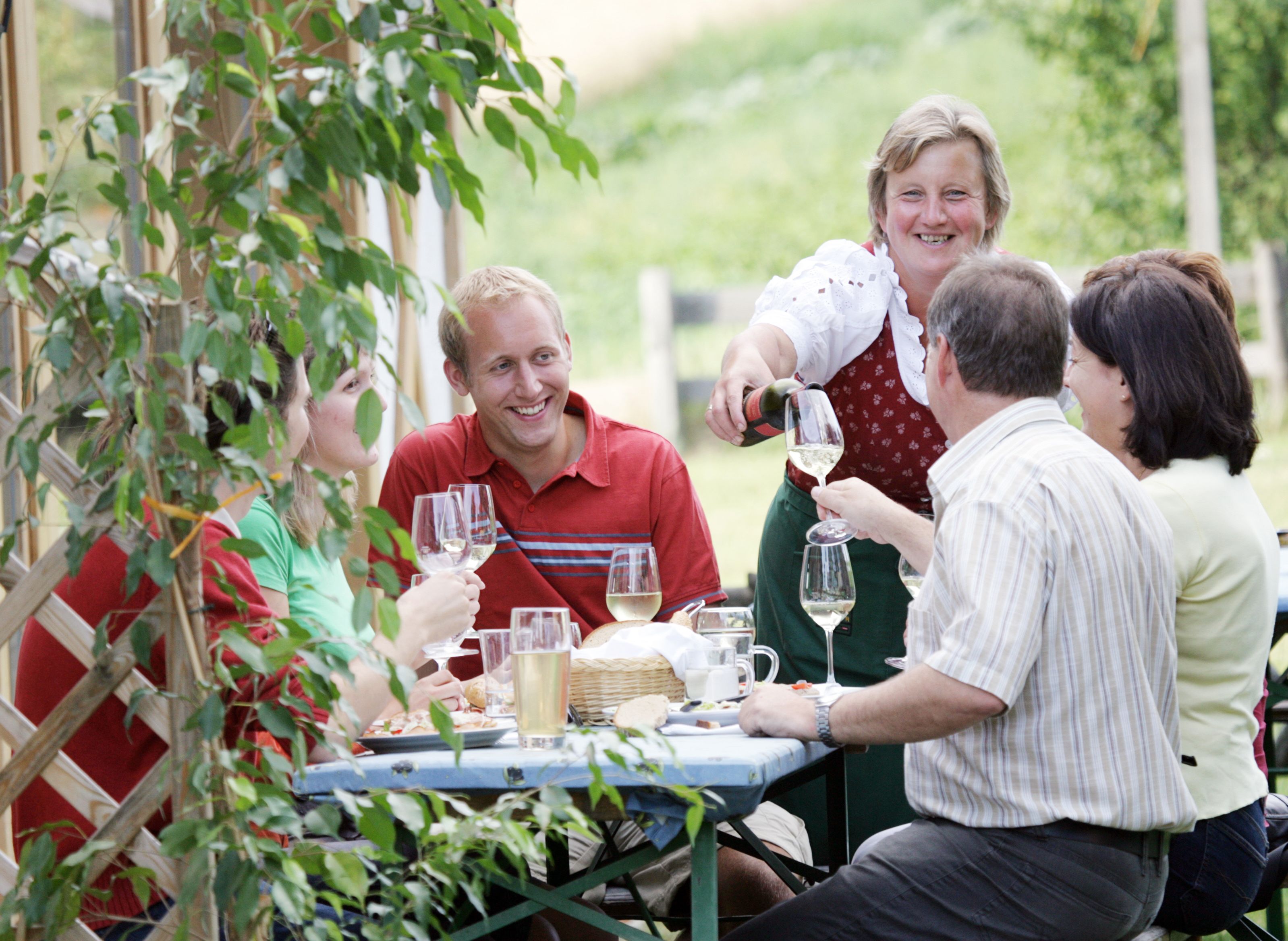 A group of people are sitting at an outdoor table enjoying wine. A woman pours wine and everyone smiles.