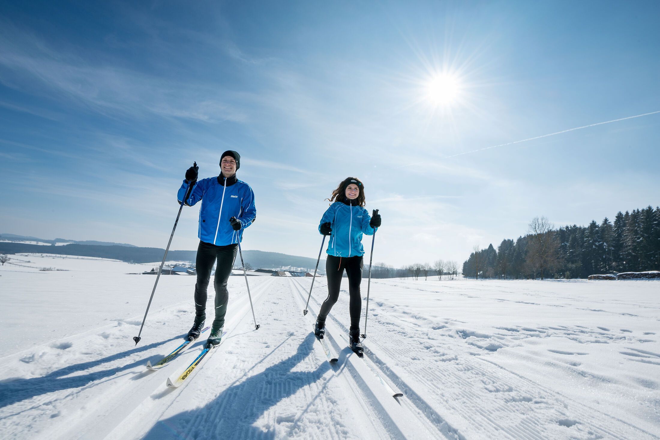Two people cross-country skiing in a snowy landscape under a blue sky and sunshine.