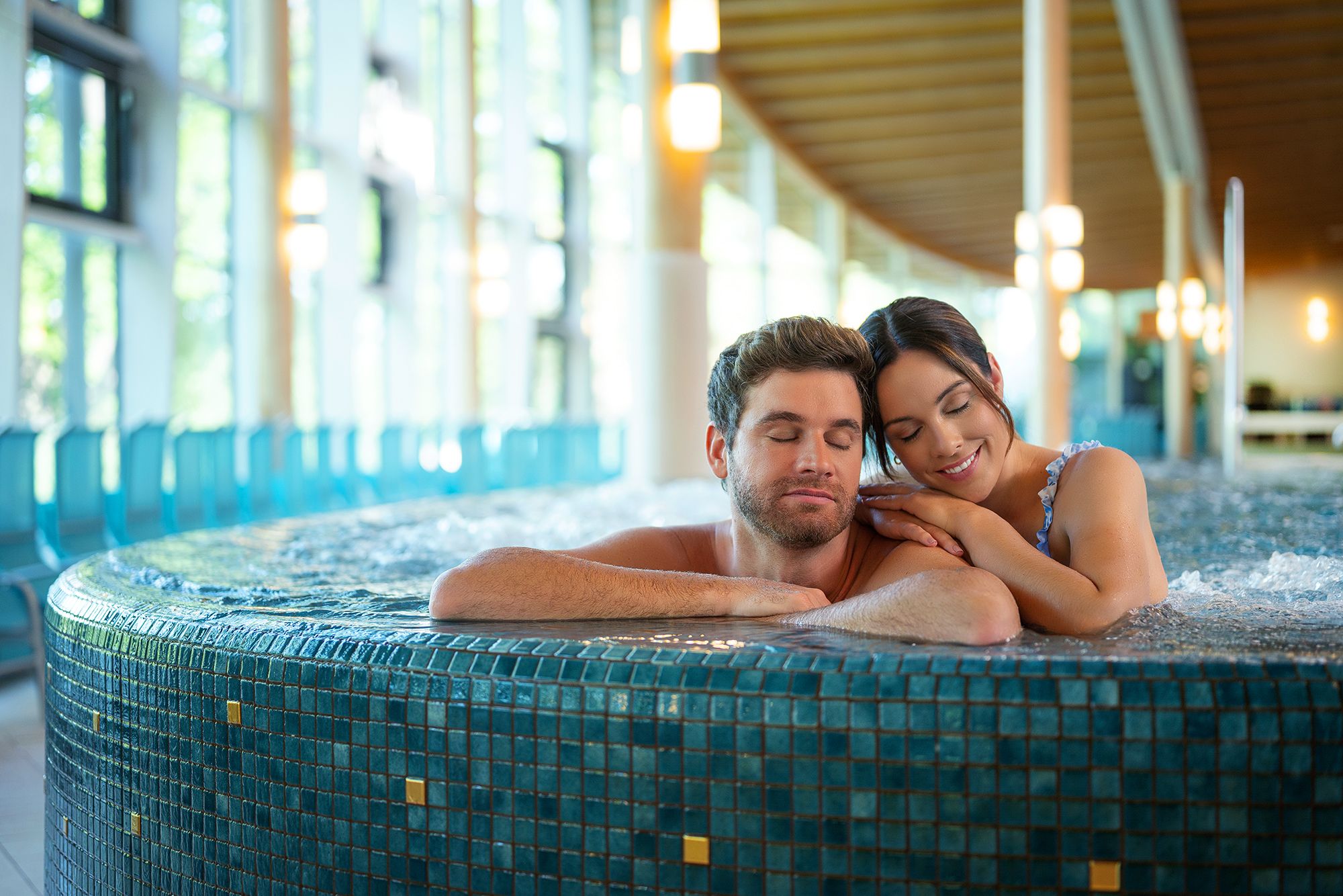 A couple relaxes in a whirlpool in a modern spa with large windows.