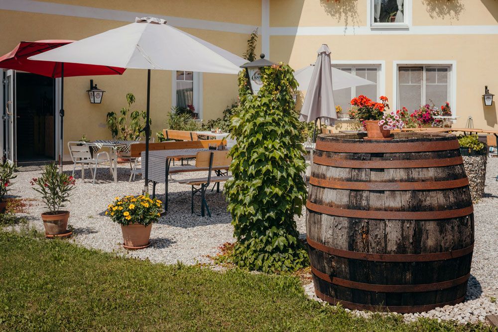 A cozy guest garden with parasols, wooden tables and a large wooden barrel, surrounded by plants and flowers.