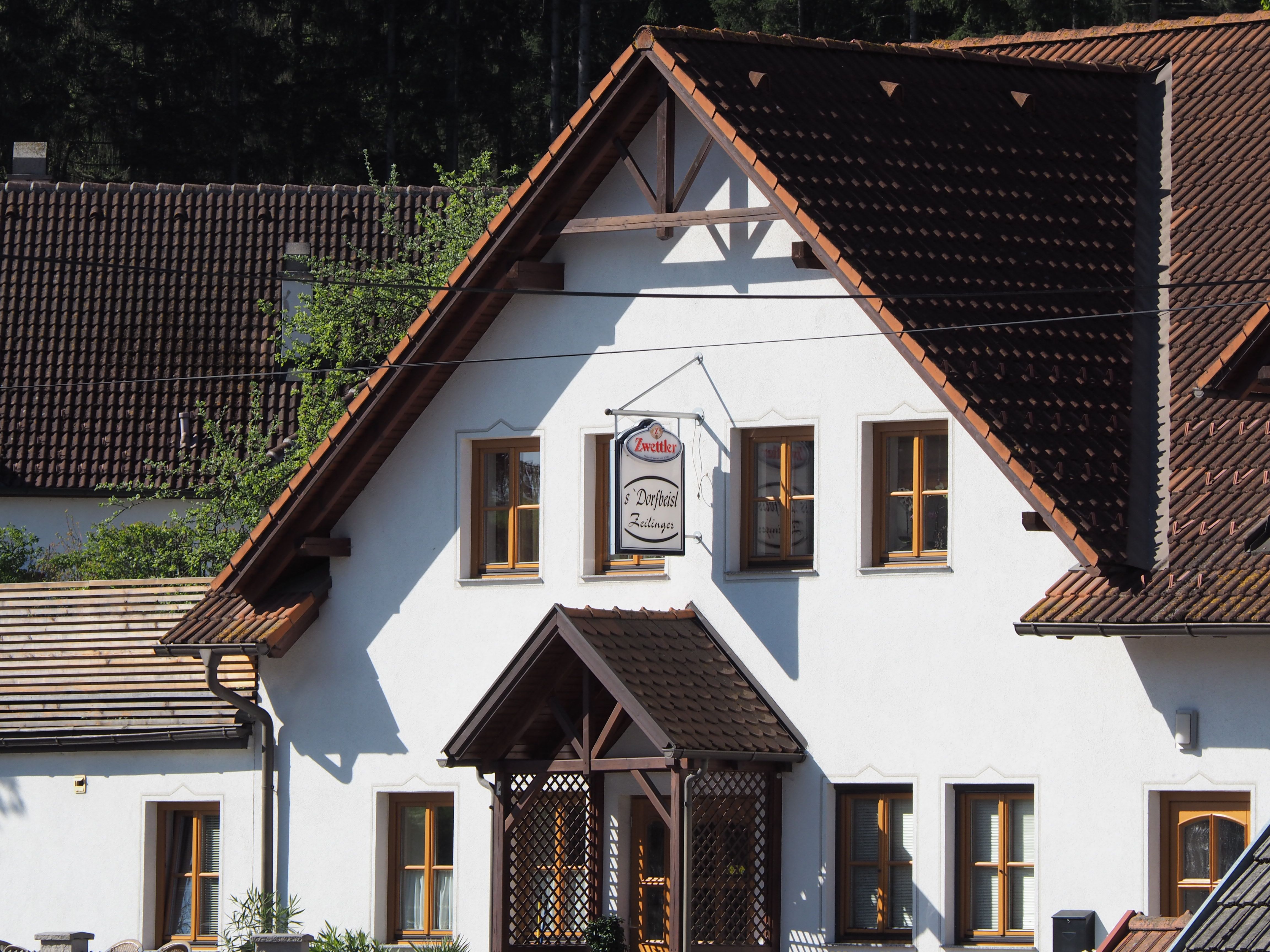 A traditional building with white walls and brown roof tiles. A sign with the inscription 'Zwettler Dorfwirt Zeilinger' hangs on the façade.