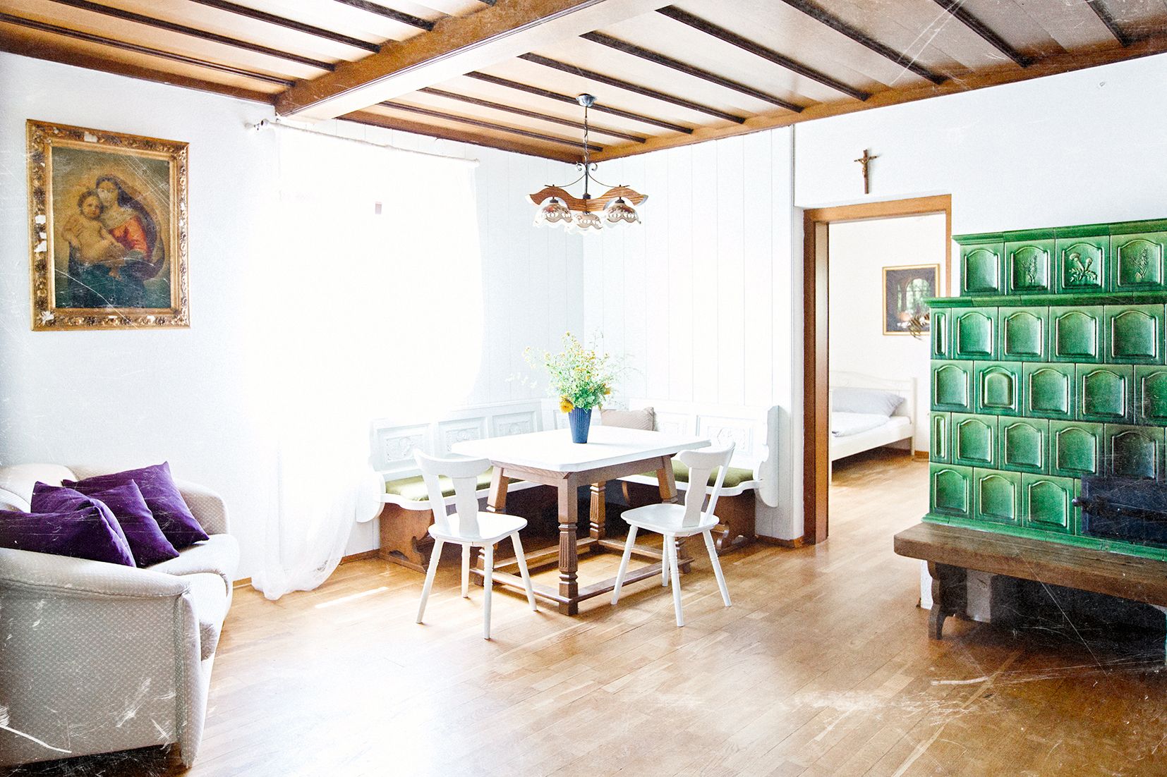 A bright living room with a wooden-beam ceiling, a green tiled stove, a dining table with chairs, and a sofa. A painting hangs on the wall.
