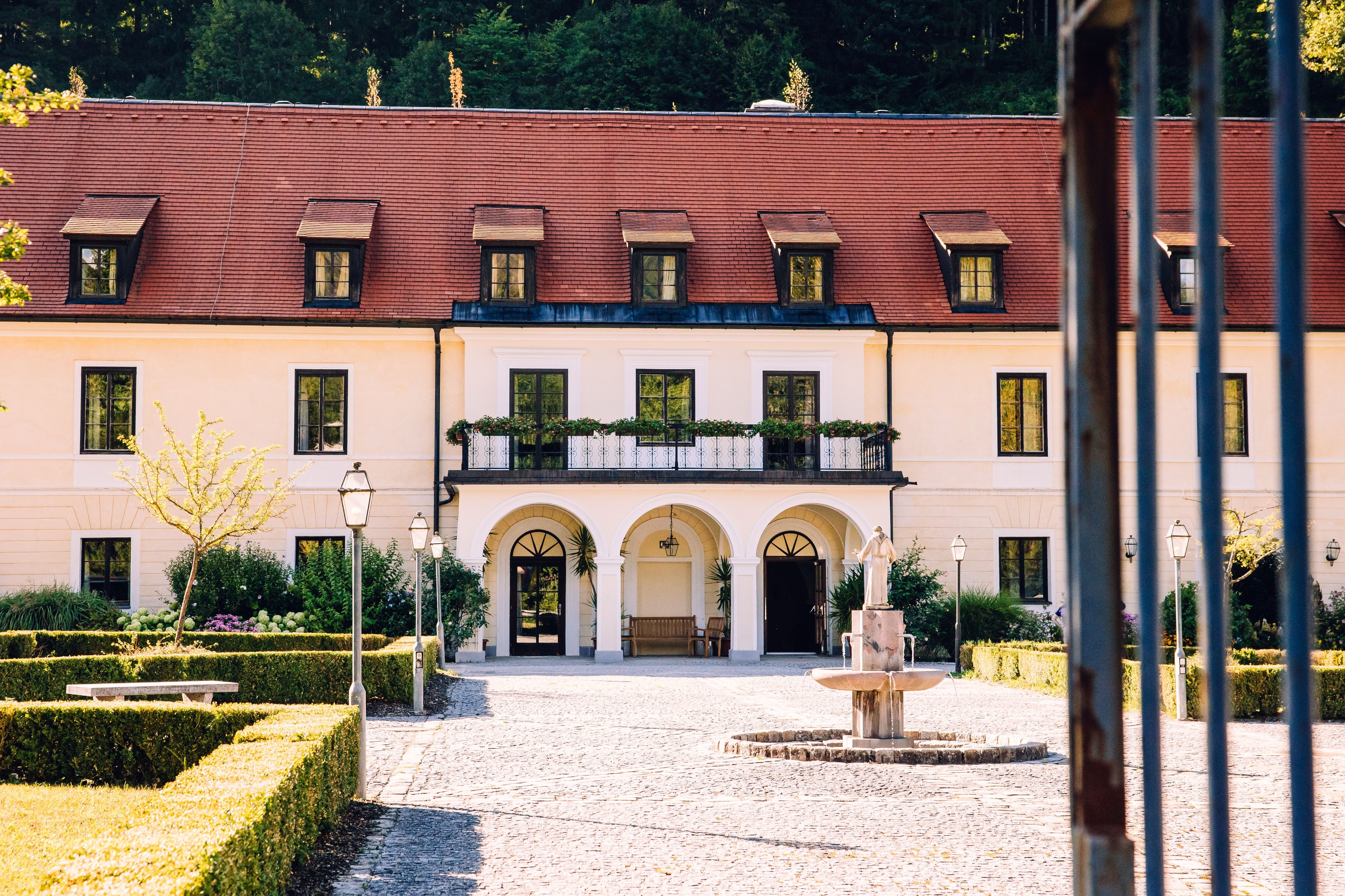 The courtyard of the Gaming Charterhouse, with its red roof, fountain, and well-trimmed hedges; in the foreground, a gate with bars.