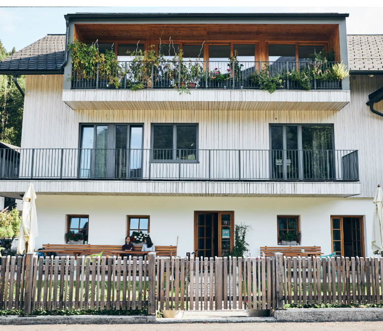 A modern three-storey house with wooden balconies and plants. Two people are sitting on a bench behind a wooden fence in front of the house.