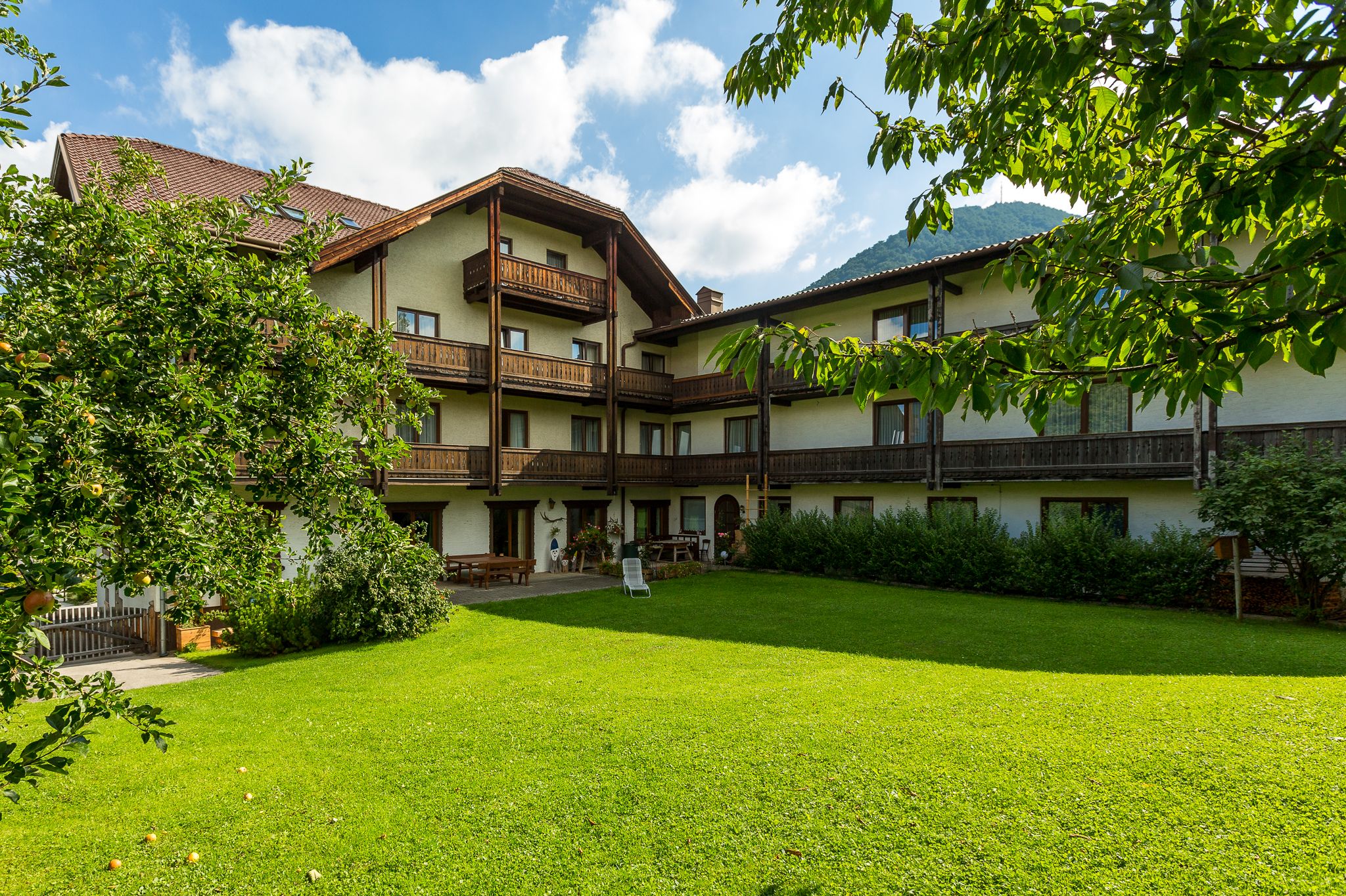 A traditional hotel with wooden balconies and a well-tended garden in the foreground, surrounded by trees and mountains in the background.