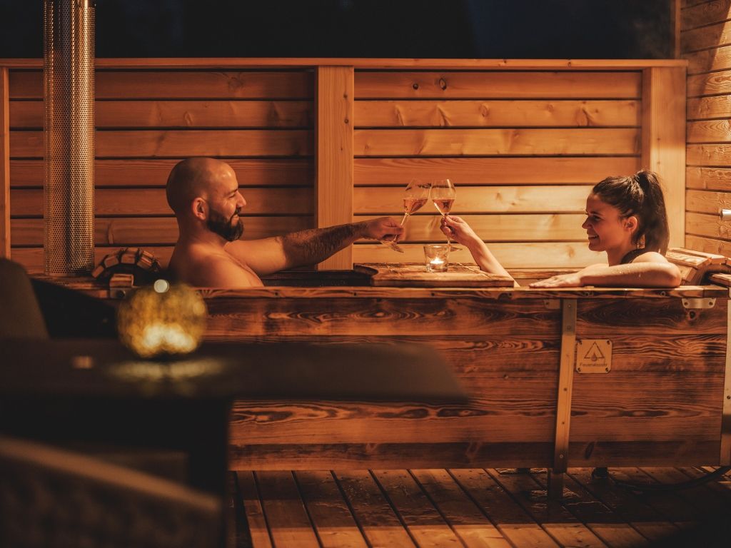 Two people in a wooden bathtub outdoors, clinking glasses of wine.
