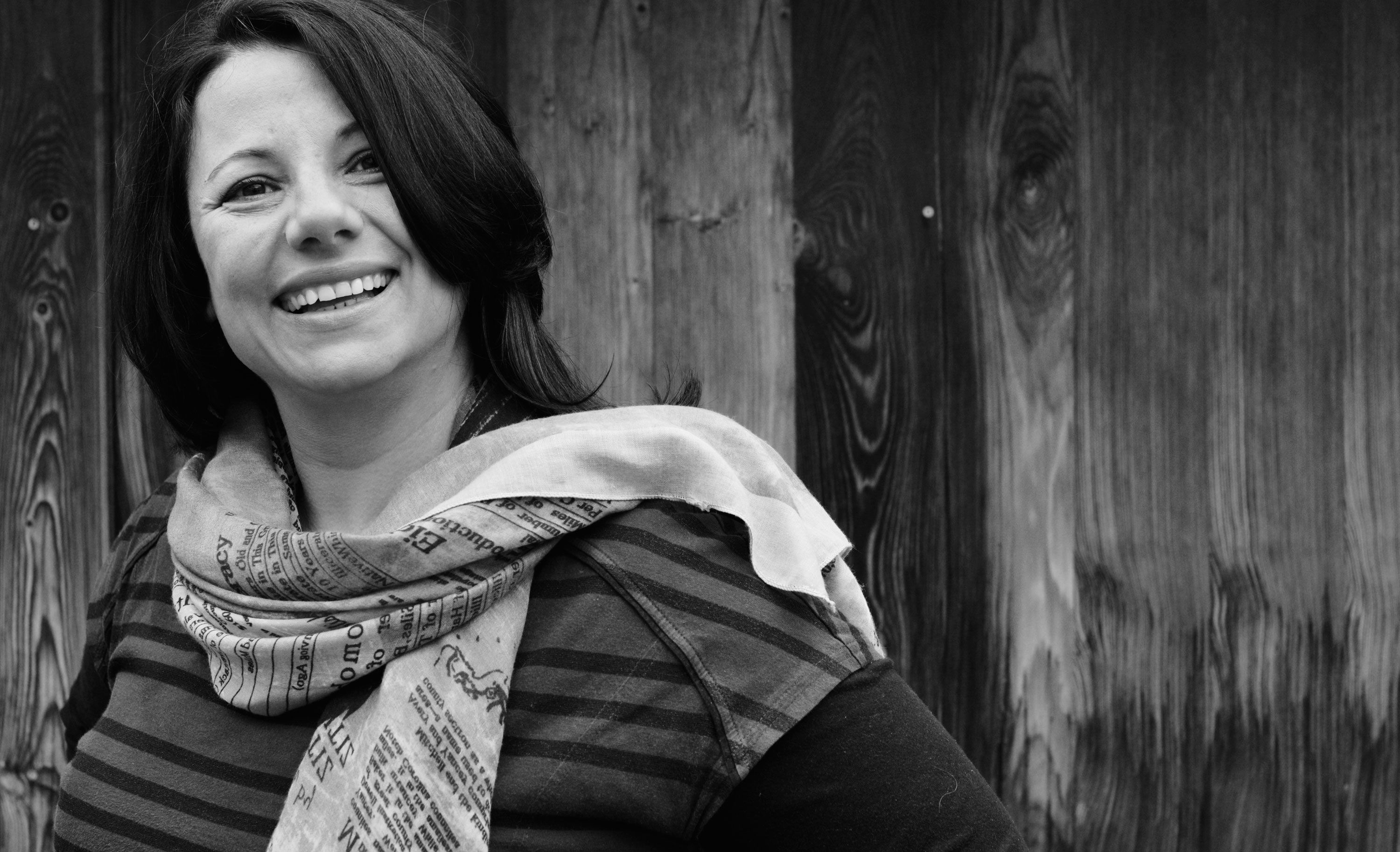 Black and white photo of a smiling woman with a scarf in front of a wooden wall.