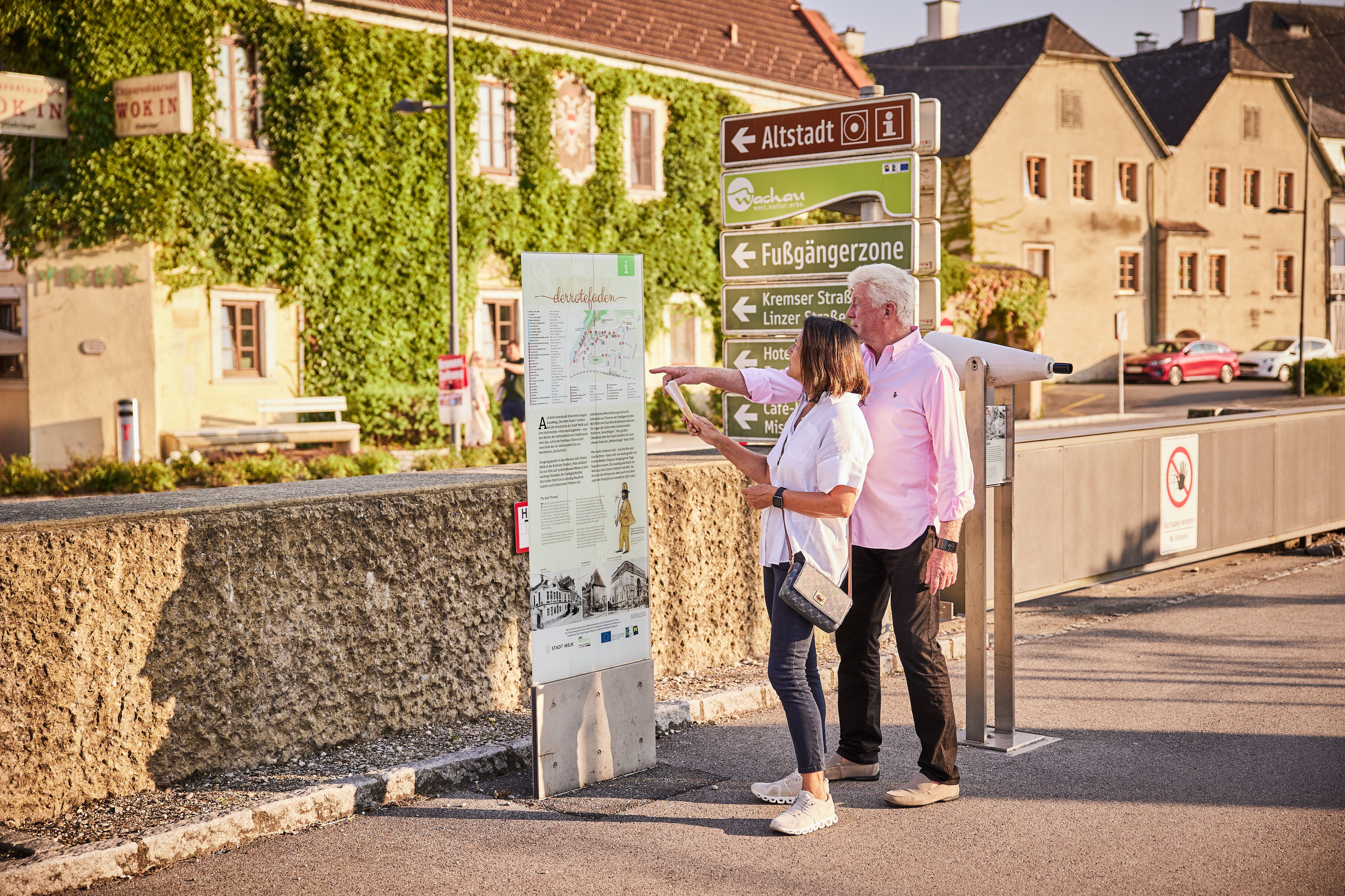 An elderly couple looks at an information board on a themed trail in a town with historic buildings in the background.