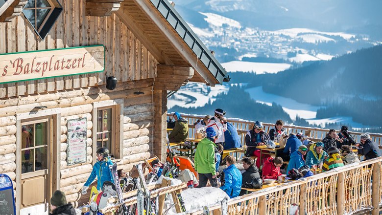 People on a sunny terrace in front of a mountain hut with a view of snow-covered mountains.