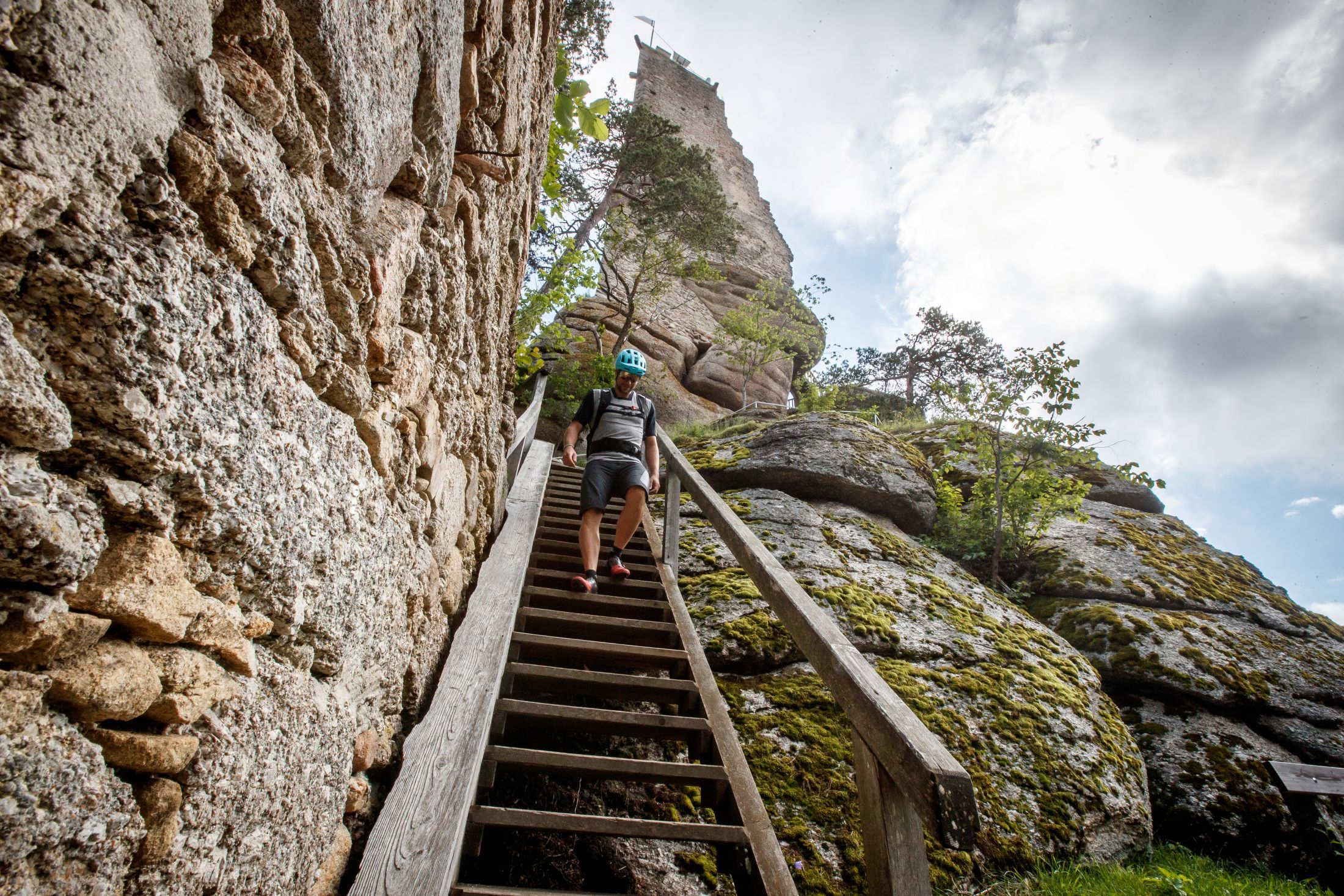 A man in a helmet walks down a wooden staircase, next to a stone castle ruin and rocks.