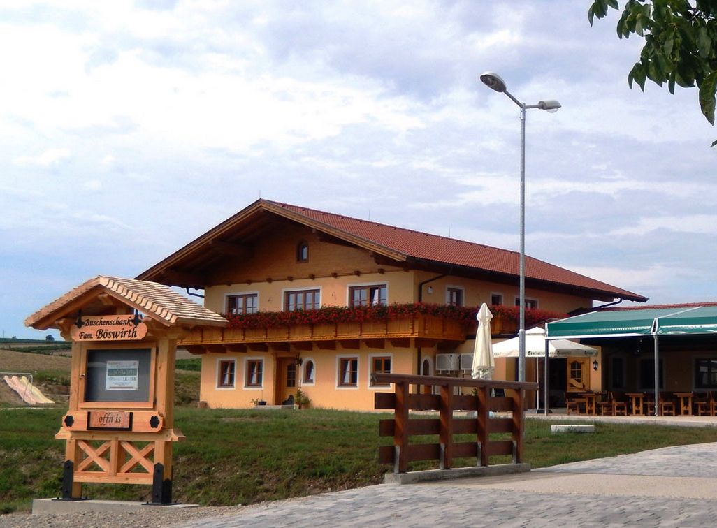 A traditional inn with a wooden balcony and floral decorations, next to a sign reading 'Buschenschank Fam. Böswirth'.