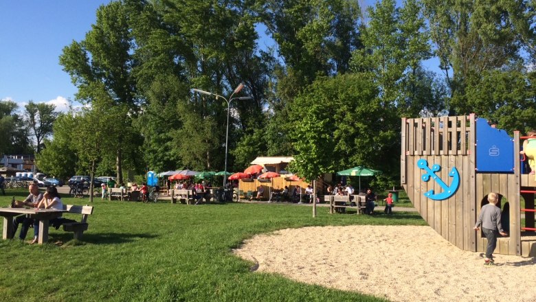 Playground with wooden climbing frame and snack area in the background, surrounded by trees and meadow.