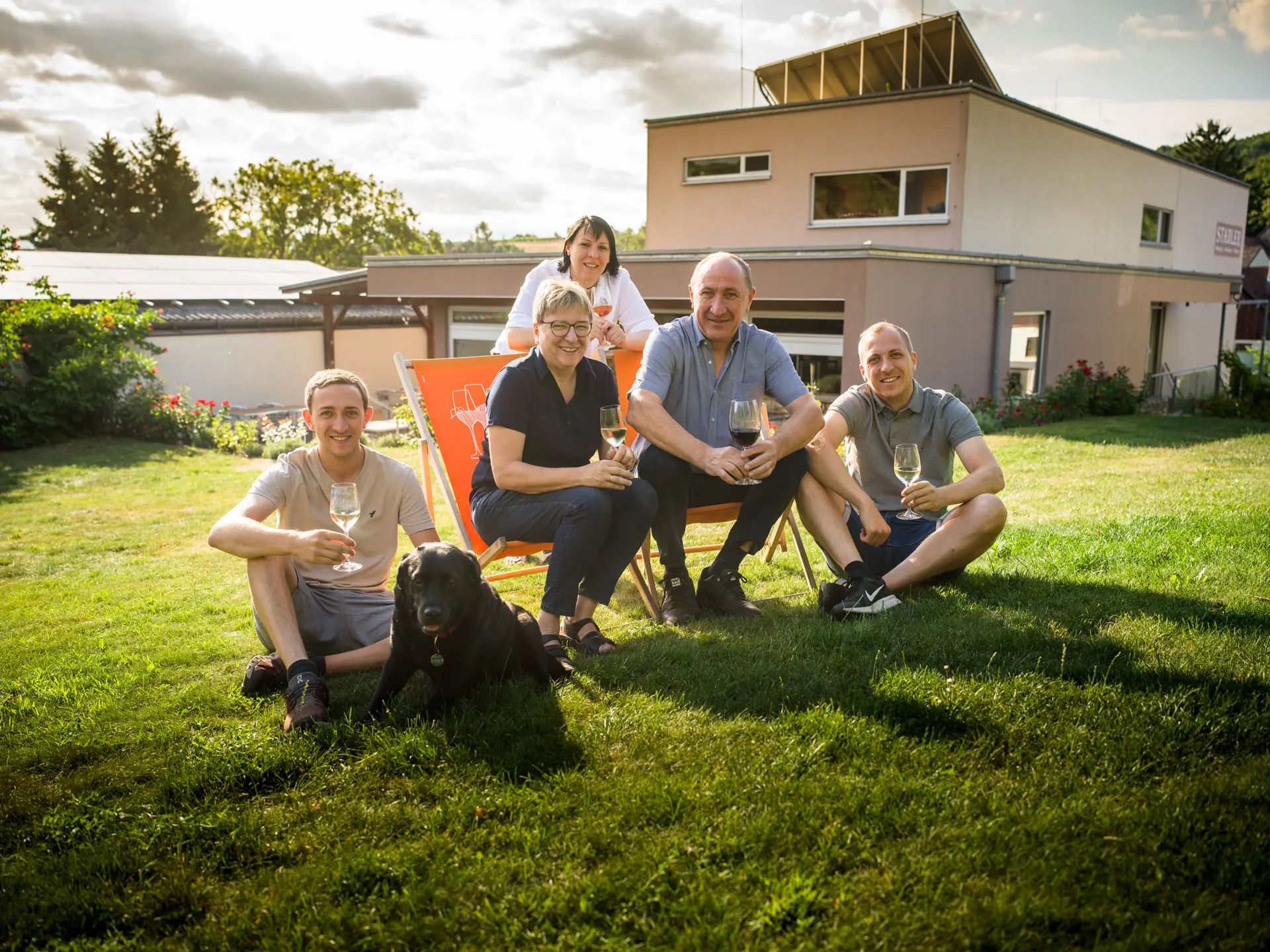 A family sits with a dog on a lawn in front of a modern house, holding glasses of wine.