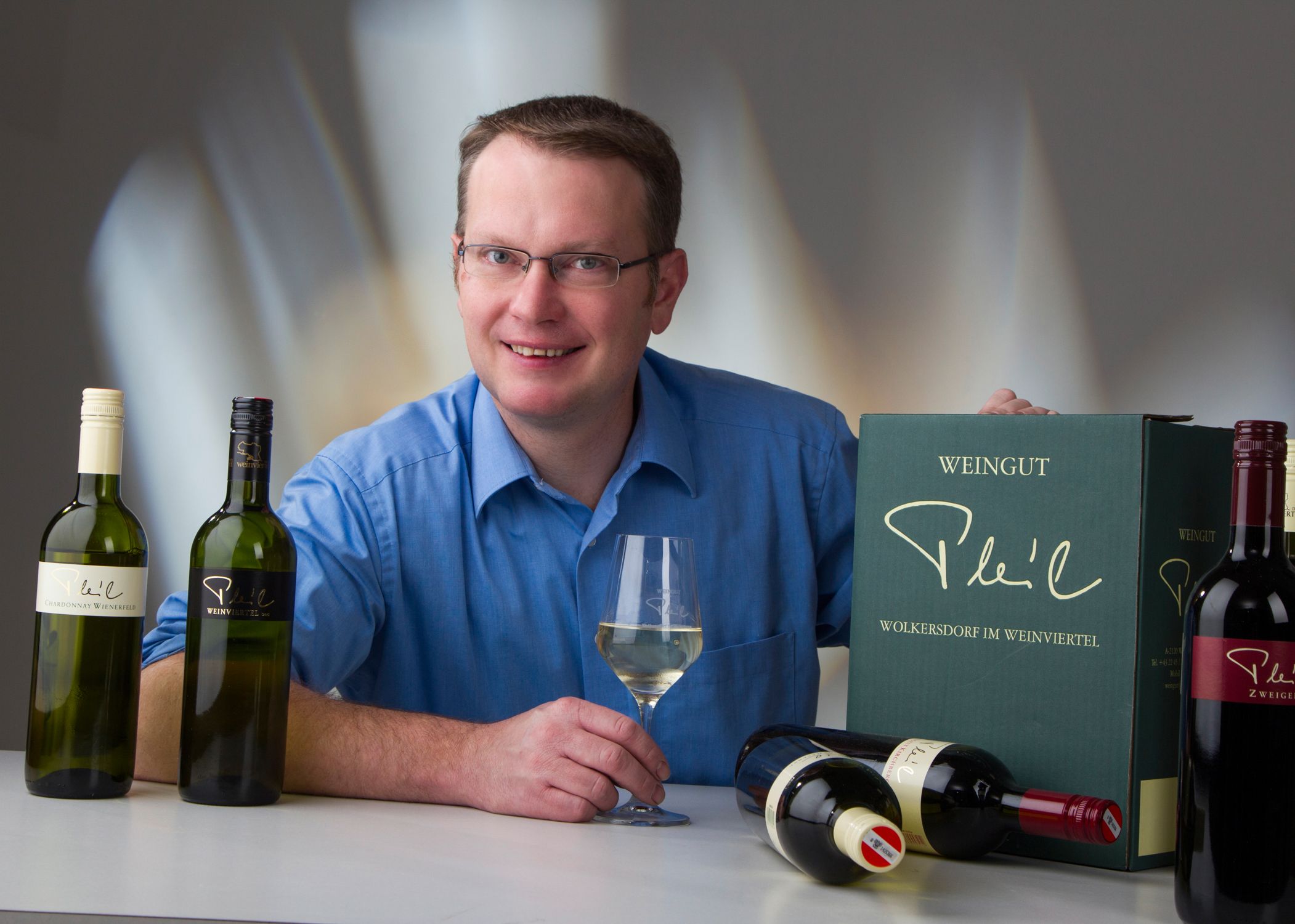 A man in a blue shirt holds a wine glass and sits in front of bottles of wine and a wine crate from the Pleil winery.
