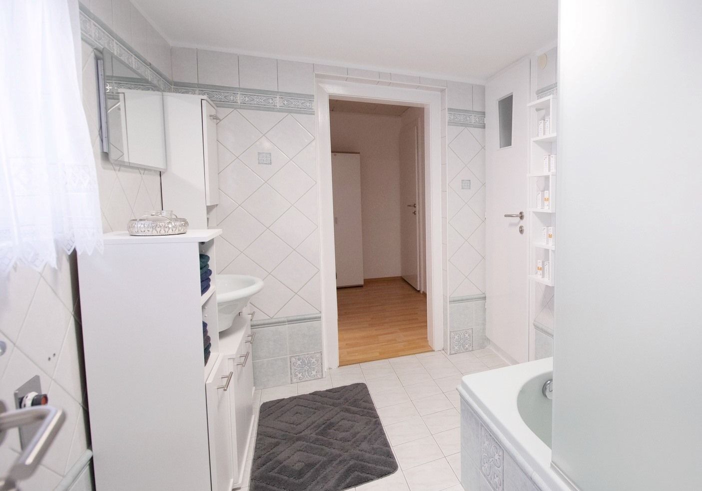 Modern bathroom with white tiles, bathtub, washbasin and cabinet. Door leads to a hallway with wooden floor.