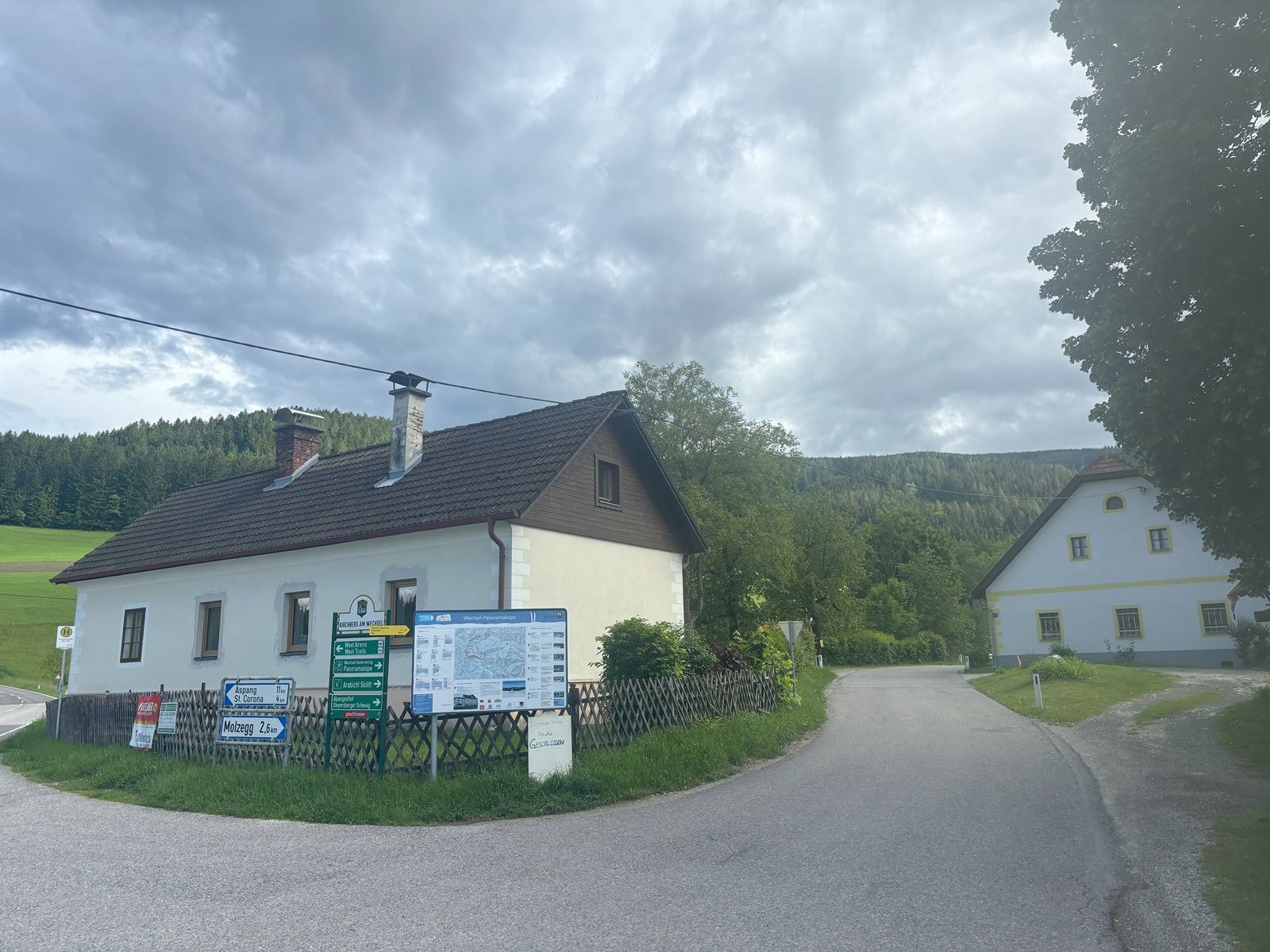 Rural road with two houses and signposts, surrounded by woods and meadows.