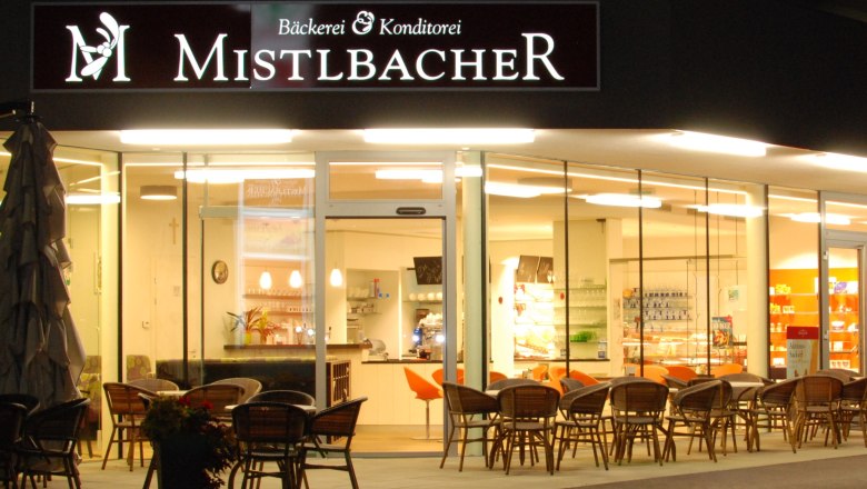 Exterior view of a café called Mistelbacher, illuminated and with outdoor seating.