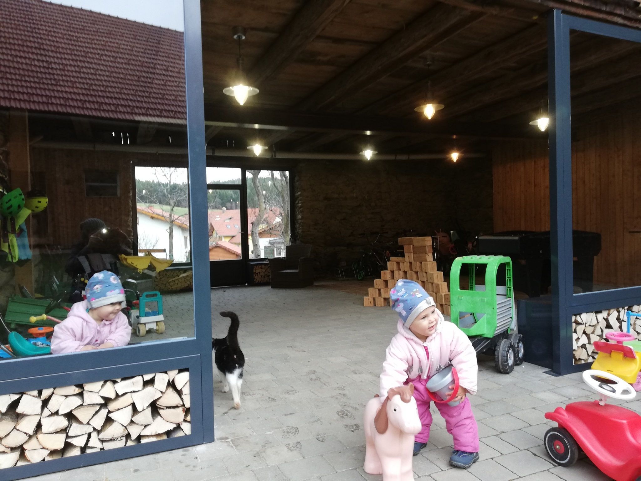 A child in winter clothes plays with a toy horse in an open room with piles of wood and toys. A cat walks past.