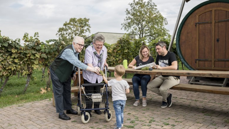 A family sits at a wooden table outside, surrounded by vines. An elderly couple is giving grapes to a child.