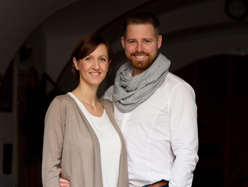 A smiling couple is standing next to each other, the man is wearing a scarf.