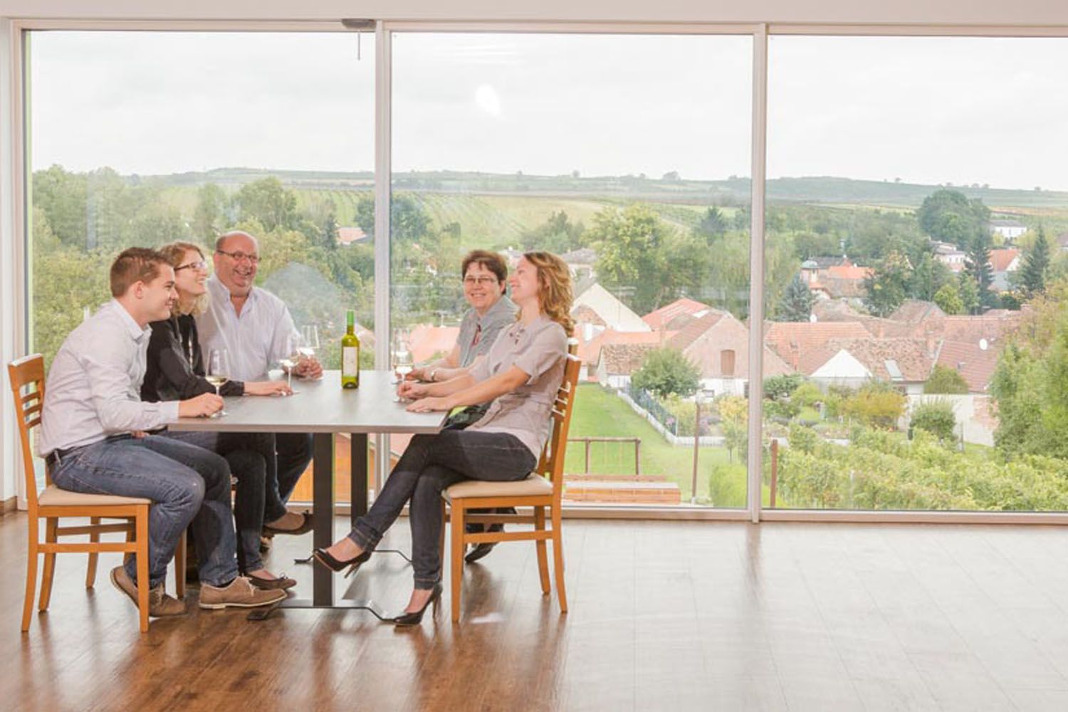 A family sits at a table with wine, in the background a landscape with houses and fields.