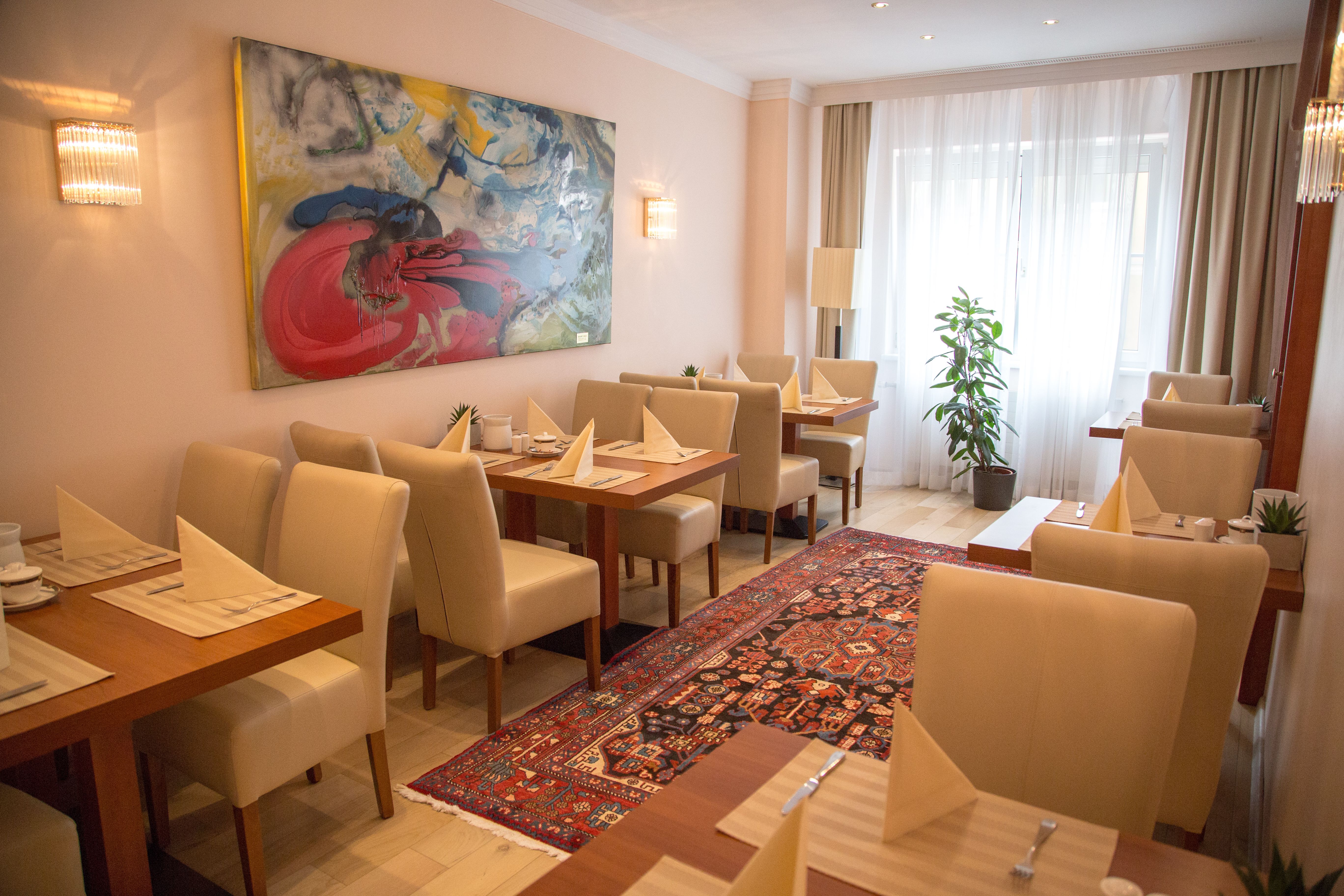 An elegant breakfast room with laid tables, beige chairs, a colorful carpet and a large abstract painting on the wall.