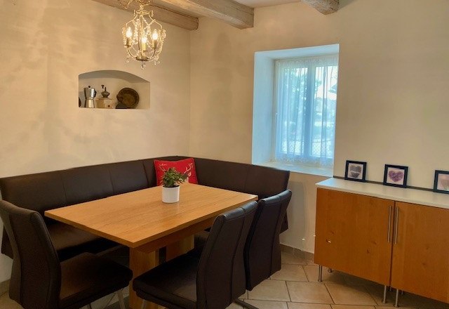 A cozy dining area with a wooden table, black chairs and a corner bench. A chandelier hangs on the wall, next to it a window and a sideboard.
