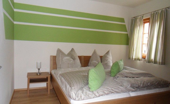 Bedroom with green and white wall decoration, double bed with green cushions, wooden floor and window with striped curtains.