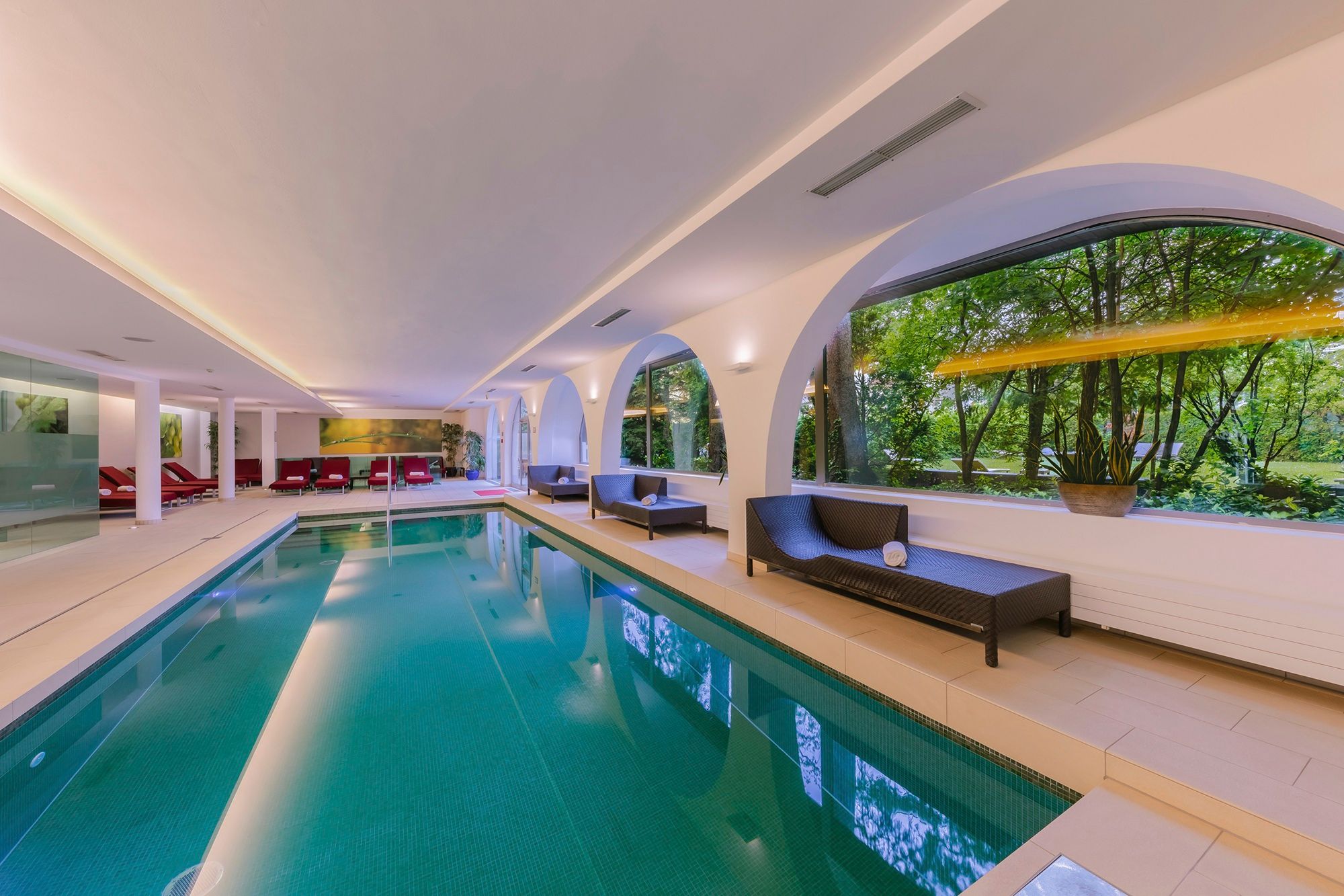 Interior view of an elegant spa area with a pool, lounge chairs, and large arched windows overlooking the garden.