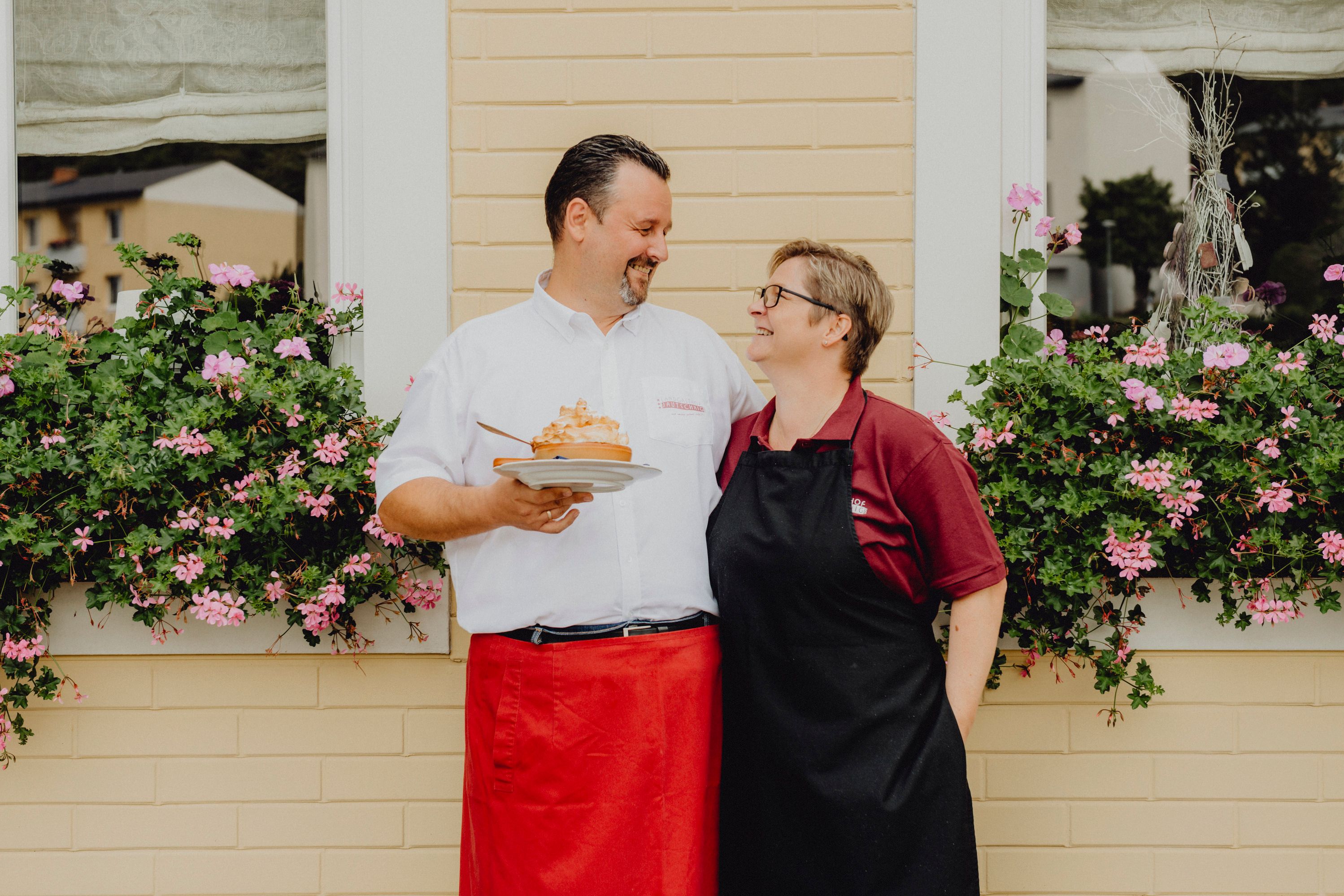 A man and a woman stand smiling in front of a window with flowers.