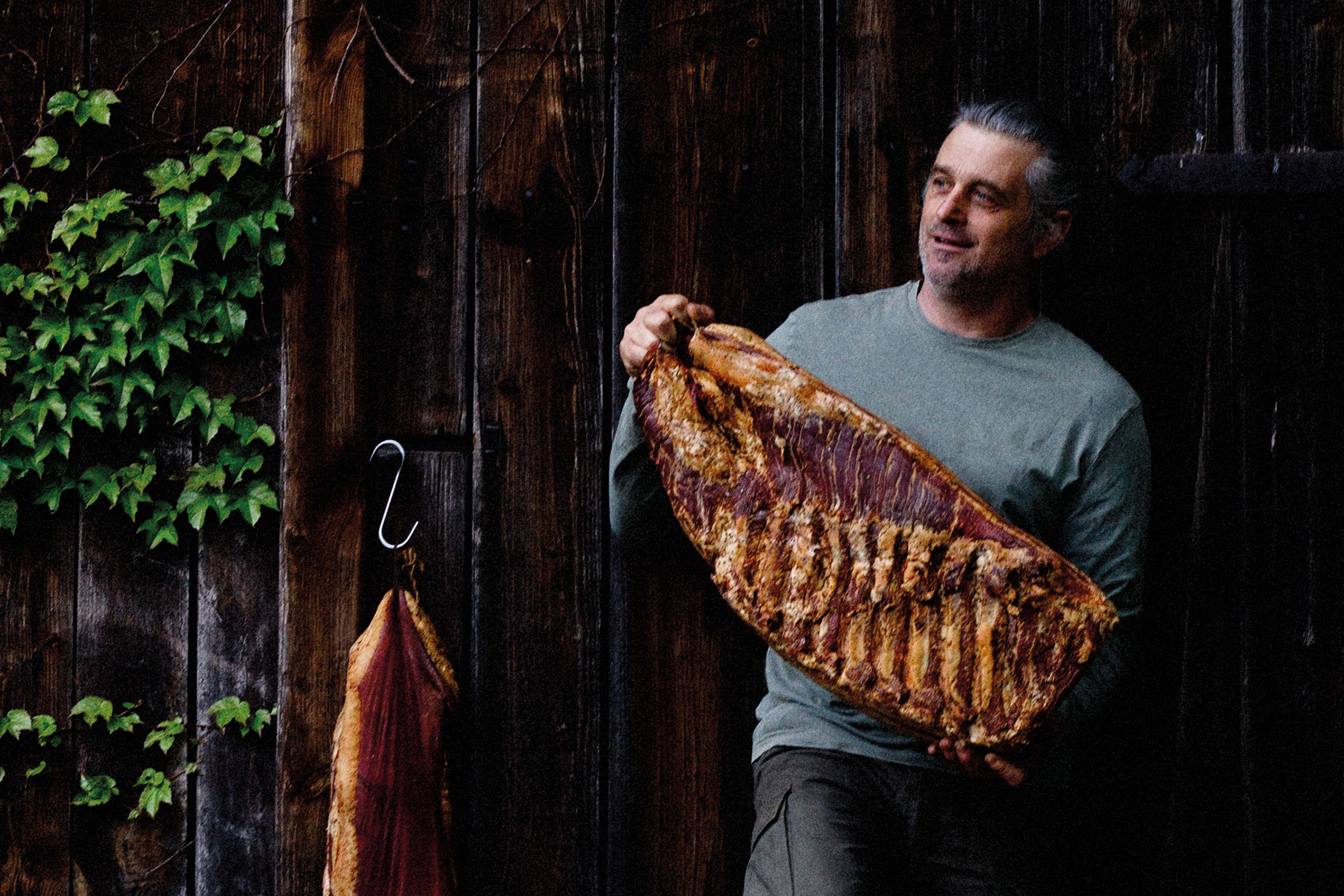 A man is holding a large piece of bacon in front of a wooden wall covered in ivy.