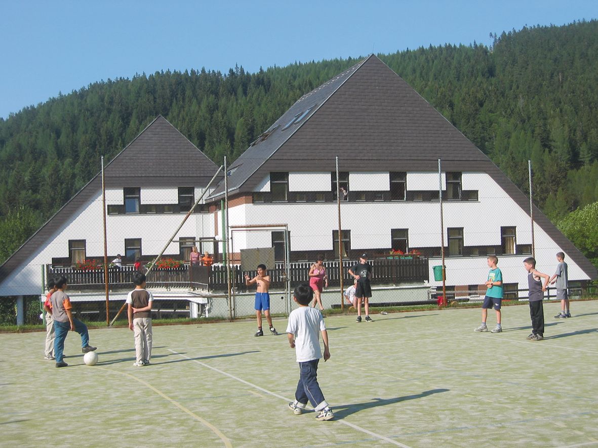 Children play soccer on a sports field in front of a hotel in a wooded area.
