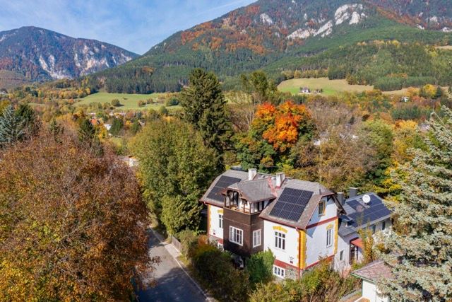 Aerial view of a country house with solar panels on the roof, surrounded by autumnal trees and mountains in the background.
