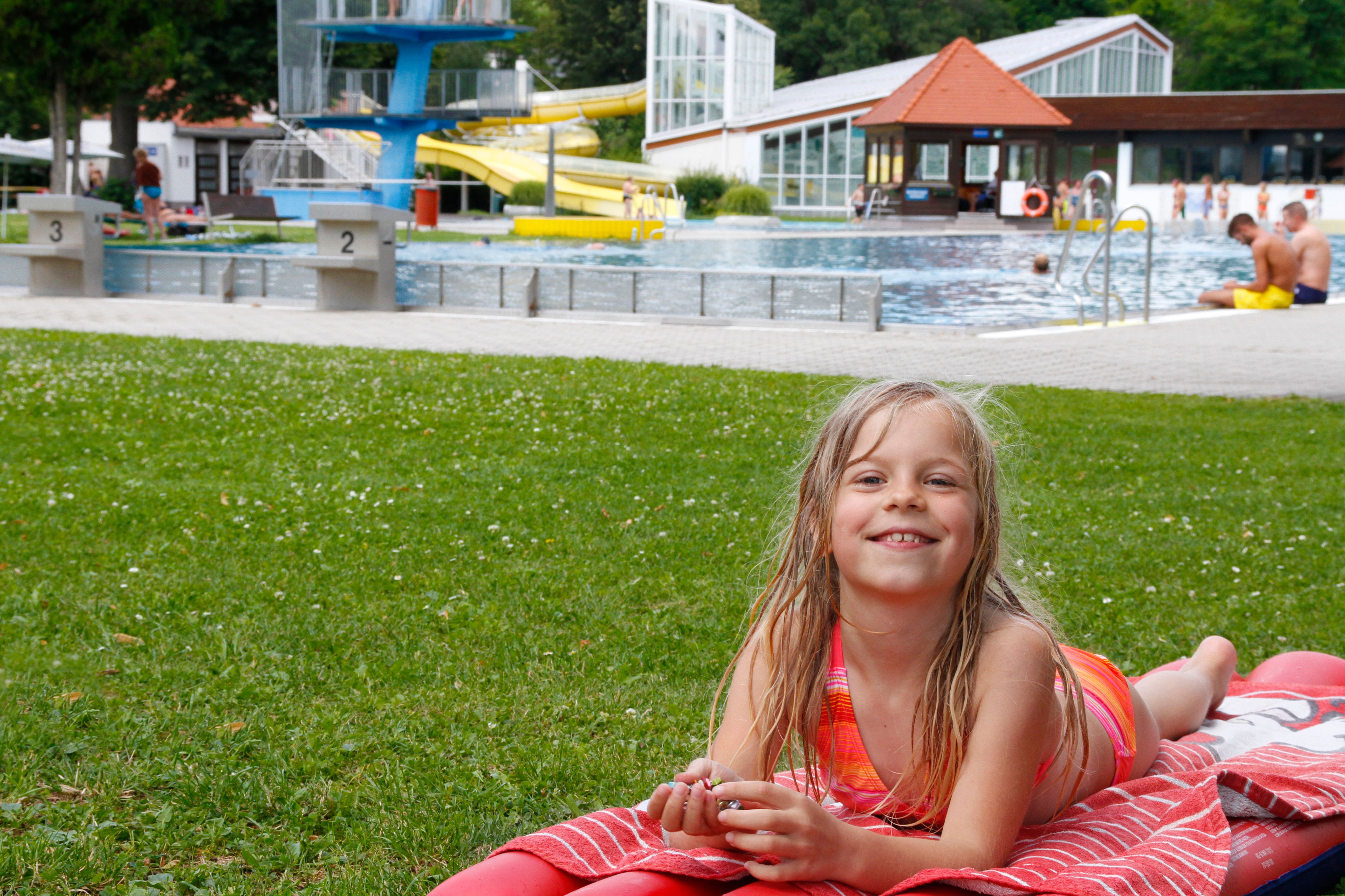 A girl lies on a red blanket in the grass in front of a swimming pool with slides and diving platform.