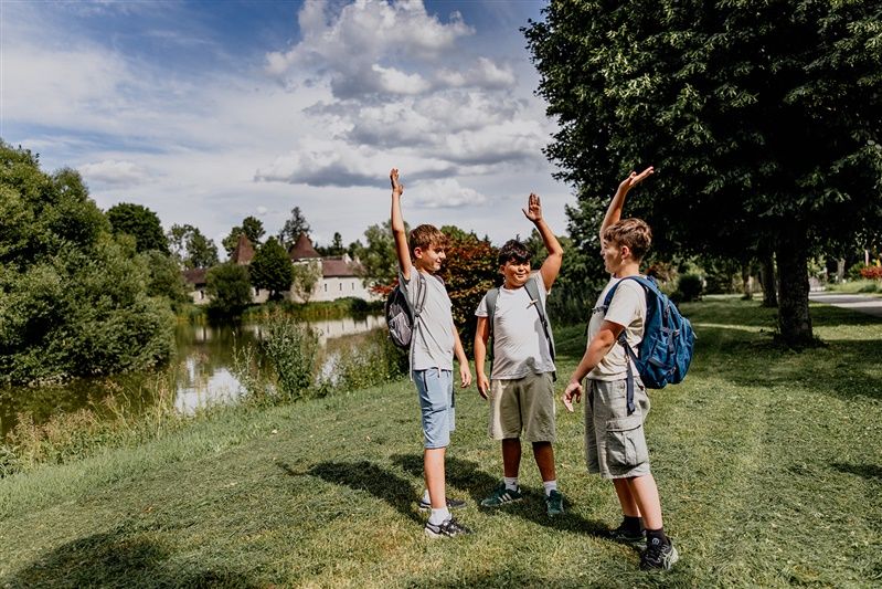 Three boys with rucksacks stand on a meadow next to a pond and raise their arms in the air.