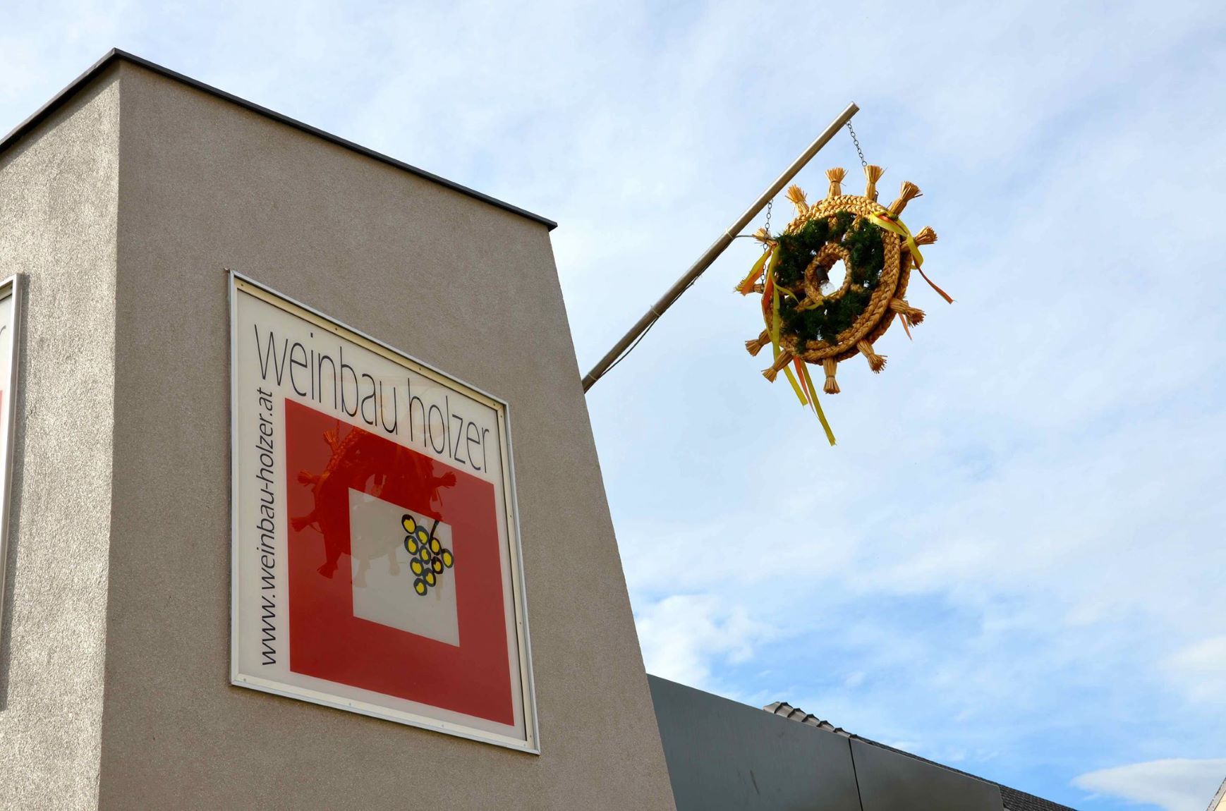 Building with sign 'Weinbau Holzer' and a decorative wreath on a pole.