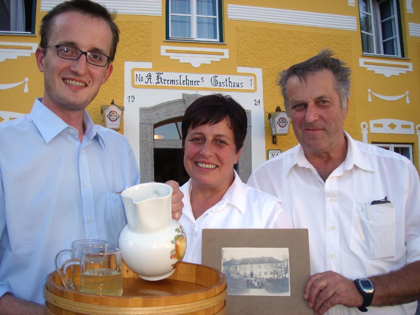 Three people in front of a yellow inn with a tray and an old photo.