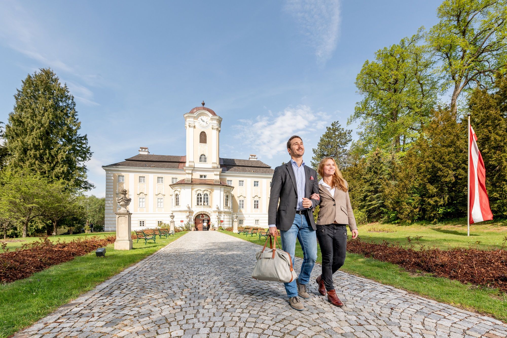 A couple walks along a cobbled path in front of the Schlosshotel Rosenau, surrounded by green trees and an Austrian flag.