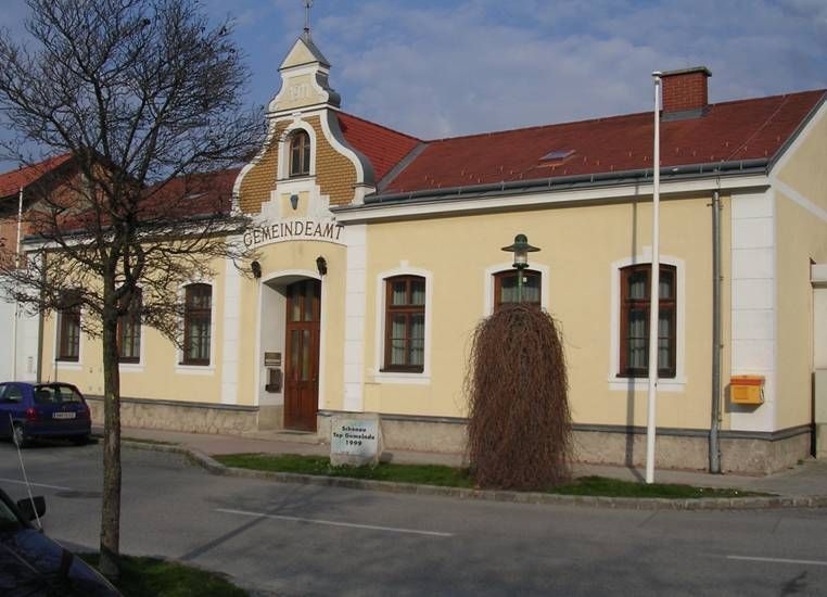 Yellow building with the inscription 'Gemeindeamt', red roof, tree and lantern in front of it.